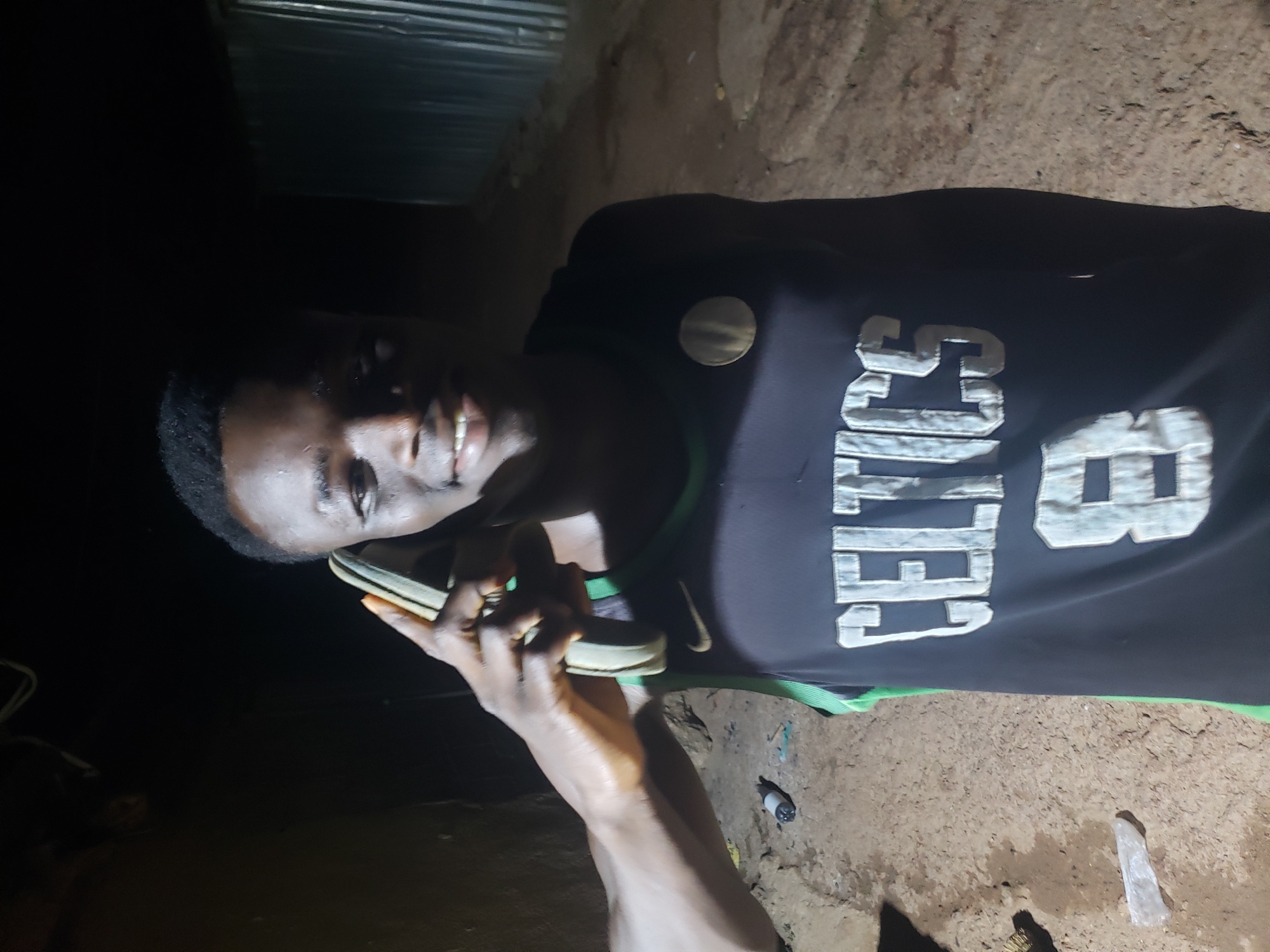 A dark-skinned male is holding a smartphone to his ear, with his left arm extended in front of him. He is wearing a dark-colored Boston Celtics jersey with a green trim. The location is dimly lit and outdoors. The ground is dirt with scattered debris. Corrugated metal sheets are visible in the background. The scene is likely Bukuru, Nigeria, though without additional context, it is difficult to corroborate it is.