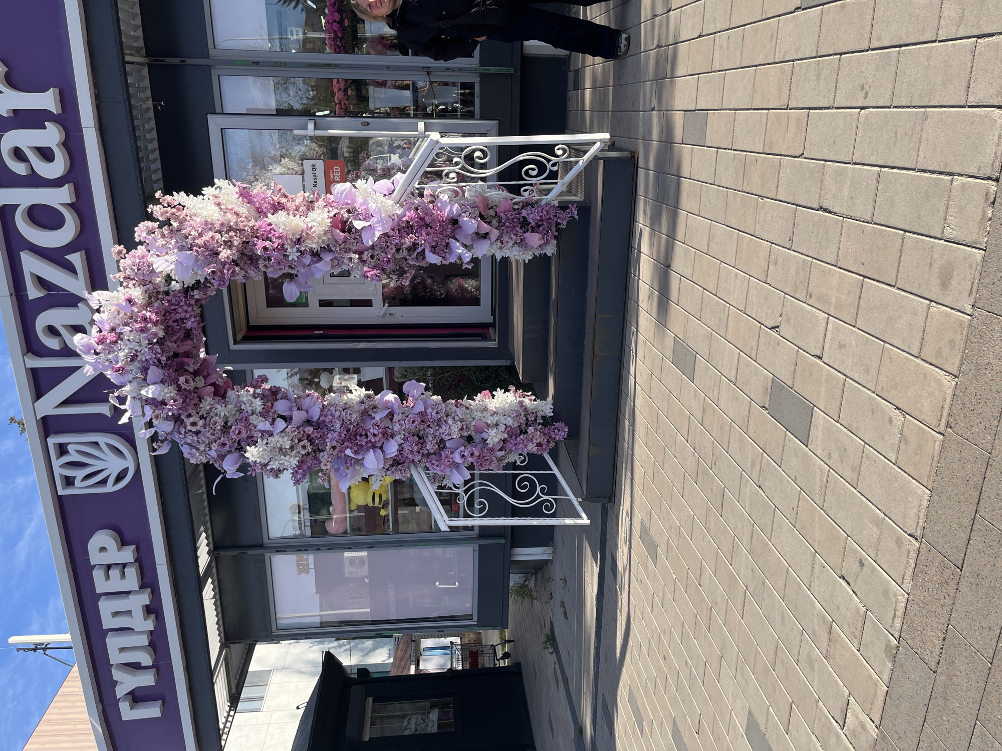 The image captures the entrance to a shop adorned with a large floral arch. The shop's signage, in purple and white, reads "ГУЛДЕР" and "Nazdar". A paved walkway leads up to a set of steps, flanked by white ornate railings, that ascend to the shop entrance. A woman stands on the right side of the entrance, partially visible, wearing dark clothing. The scene appears to be outdoors on a sunny day. The text on the signage and the presence of "Kaspi QR" on a sticker near the entrance suggest the location is in Kazakhstan, likely in a city like Astana.