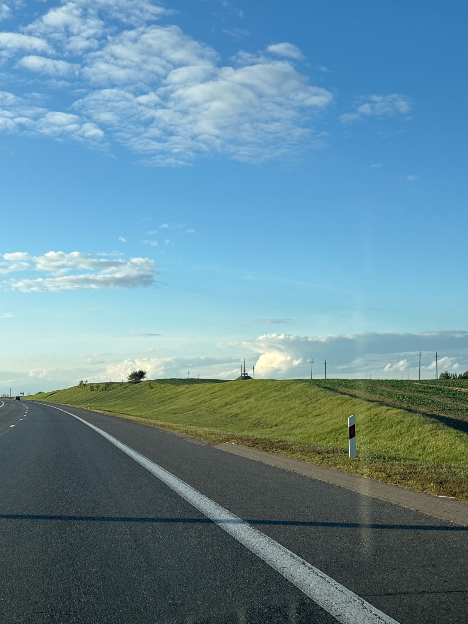 The image depicts a scenic view of a rural landscape in the city of Azyarytskaslabadski rural council, Belarus. A paved road, marked with a white line, curves through the scene, bordered by a grassy embankment on the right. Beyond the embankment, agricultural fields are visible, with rows of crops. Several utility poles are scattered across the horizon. The sky is a bright blue, adorned with fluffy white clouds. The overall atmosphere is one of tranquility and natural beauty.