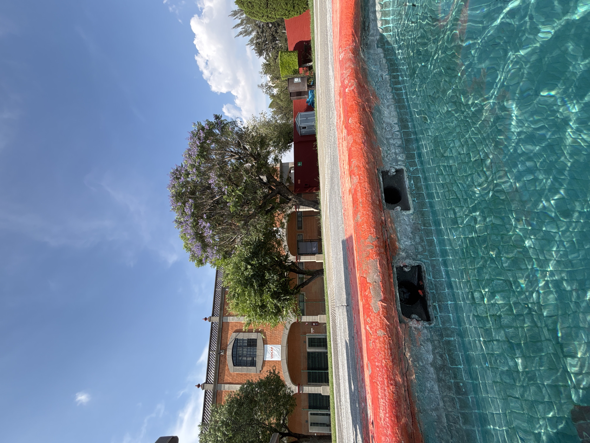 The image shows a body of water with a red-painted edge in the foreground. Adjacent to the water is a street leading to a brick building with windows and arched entrances. A tree with purple flowers partially obscures the building. The sky is blue with scattered clouds. The scene is in San Andrés Cholula, Mexico, as indicated by the "UDLAP" banner visible on the building.