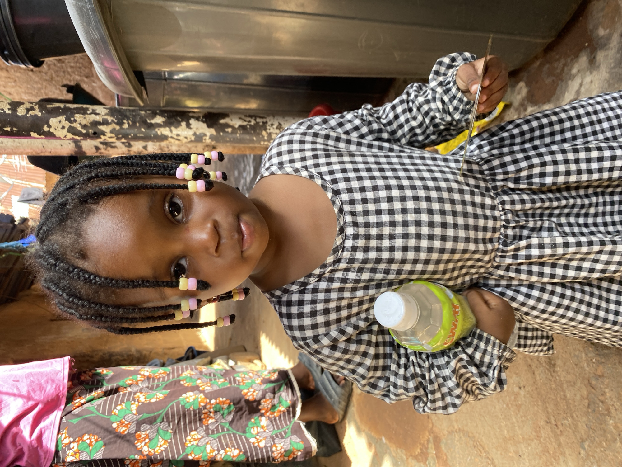 A young Black girl with braided hair adorned with pink and yellow beads is captured in a close-up, head-and-shoulders shot. She wears a black and white checkered dress with ruffled sleeves. In her right hand, she holds a thin, flat object, possibly a piece of wood or metal. Her left hand clutches a plastic water bottle with a green label. Her gaze is directed slightly upwards and to her left, with a neutral expression. The background is somewhat blurred, showing earthy tones and hints of what appears to be an outdoor setting, possibly under some structure. The lighting suggests it is daytime, and the environment appears to be simple and rustic. The scene is set in Jos, Nigeria. No visible text is legible on the water bottle or elsewhere in the frame.