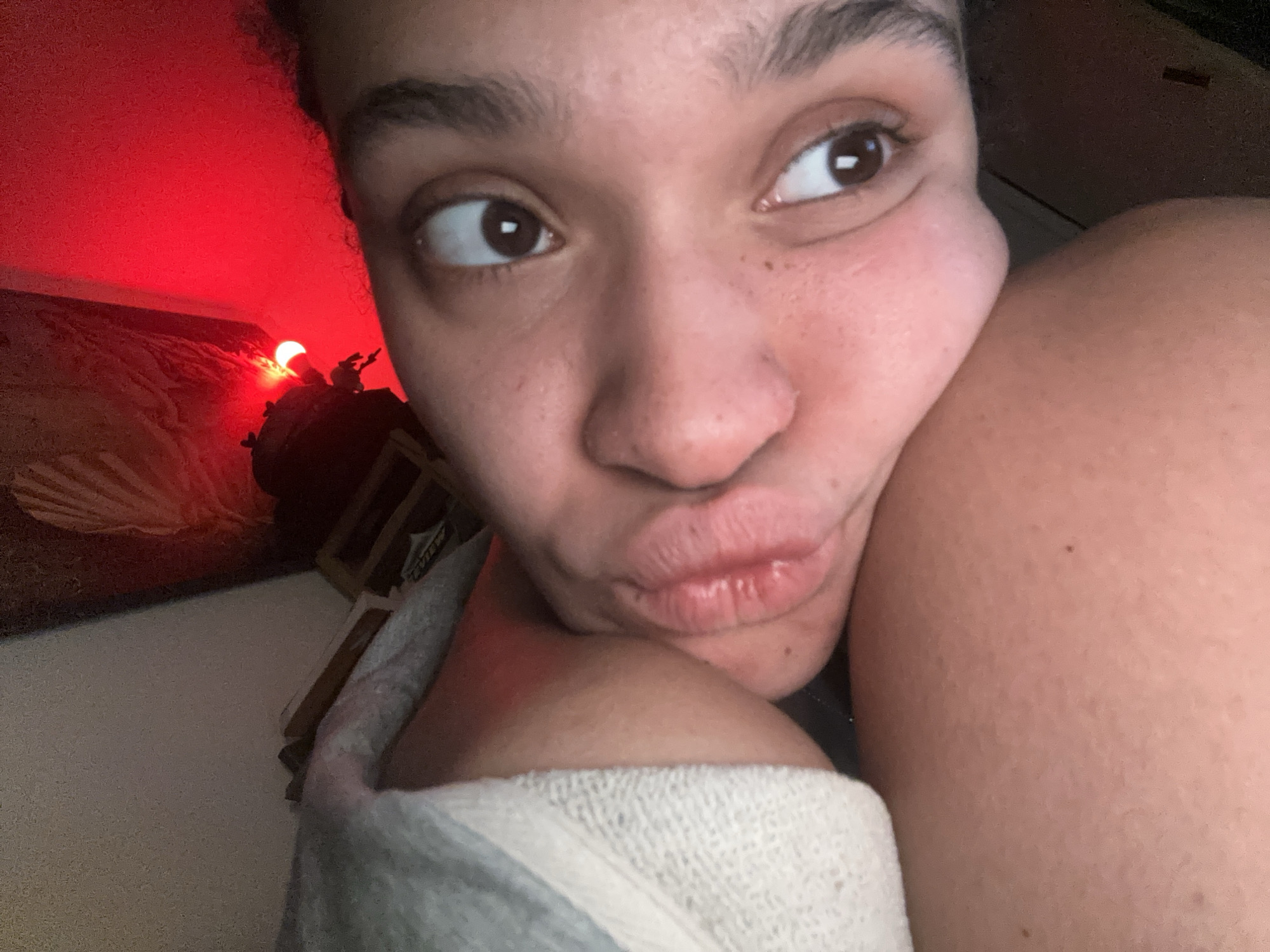 A selfie taken indoors shows a woman with brown eyes and skin pursing her lips. A partially visible white towel is wrapped around her shoulder. In the background, a red light illuminates part of a seashell art piece and a small decoration with a light. The location is undeterminable.