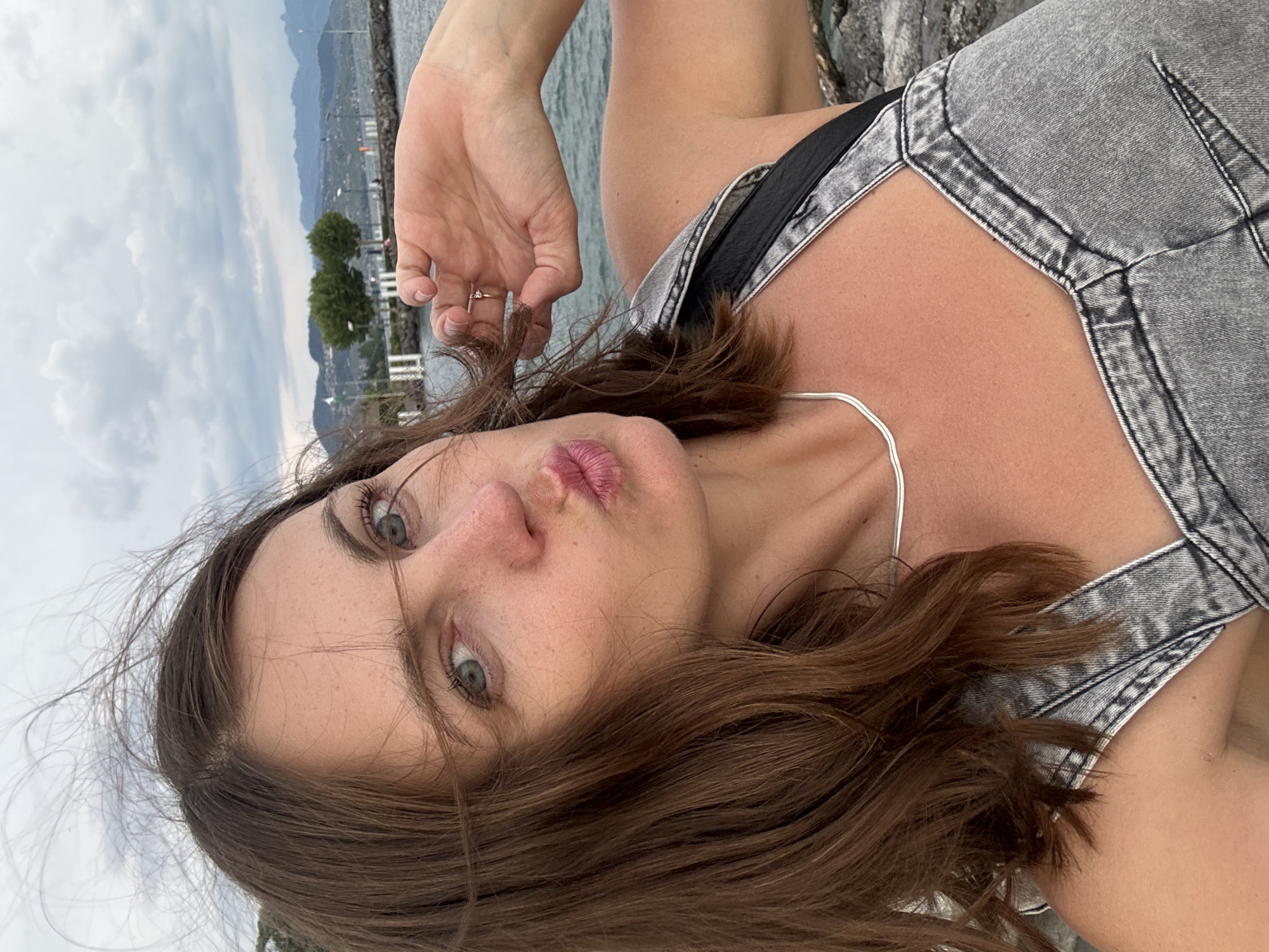 The image is a close-up selfie of a woman with brown hair, green eyes, and freckles, leaning forward and puckering her lips. She is wearing a denim overall with a black strap and a silver necklace. Her right hand is raised to her hair, and she is wearing a ring on her ring finger. The background shows a body of water, mountains in the distance, and a cloudy sky. This appears to be a scenic location, likely near the shores of Lake Geneva in Lausanne, Switzerland, given the characteristic landscape. The overall mood is casual and playful.