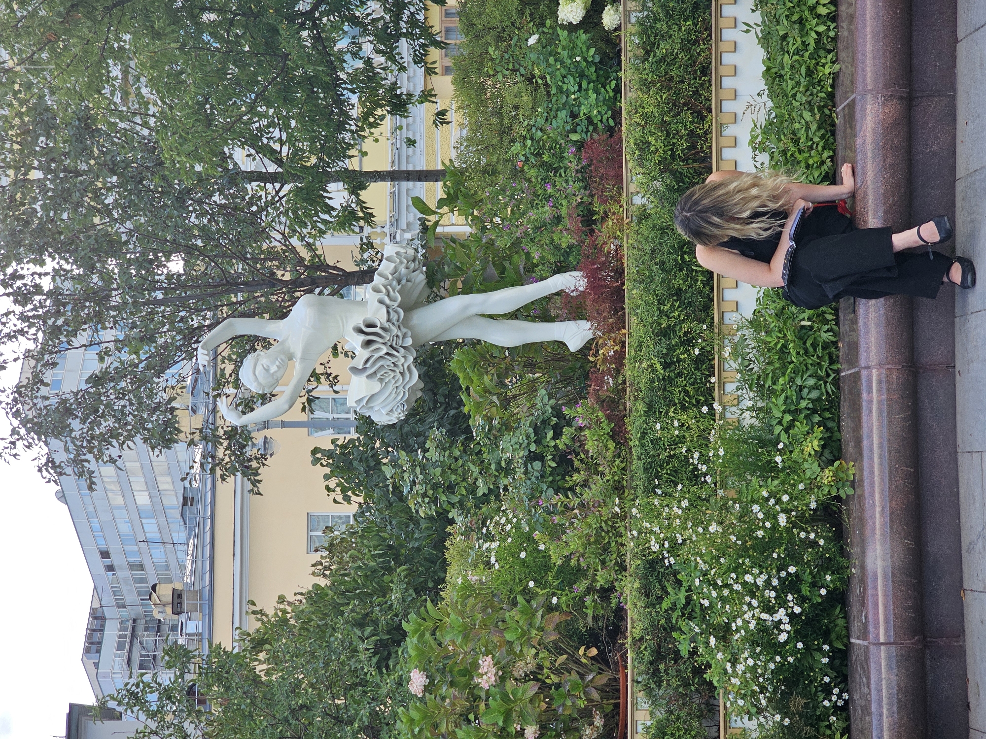 In a city park, a white ballerina statue stands gracefully amidst lush greenery and colorful flowers. In the foreground, a woman with blonde hair, wearing a black tank top and black pants, sits on a stone bench. Her head is bowed, and she appears to be reading a book. The background features a building with a modern architectural style, characterized by large glass windows.
