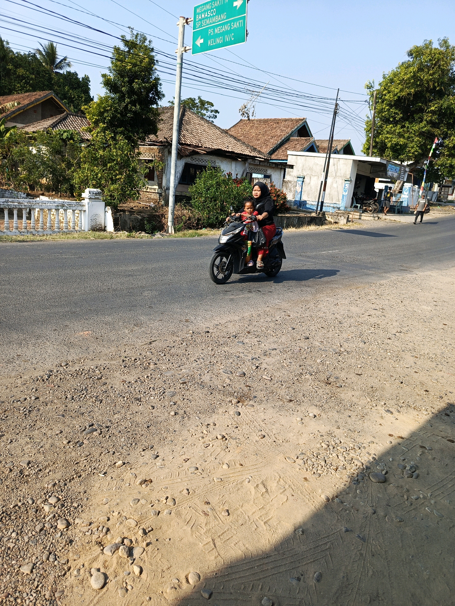 A mother wearing a black headscarf and red pants rides a black scooter with her young child in front of her. The child, wearing a red shirt, is sitting in a child seat attached to the front of the scooter. The scooter is traveling on an asphalt road. On the left side of the road is a white decorative fence and some houses with red tile roofs. On the right side of the road is a blue and white building, a street sign, and some trees. The street sign indicates directions to "MEGANG SAKTI II", "BAMASCO SP. SEMAMBANG", and "PS MEGANG SAKTI KELINGI IV/ G". The scene appears to be in a rural or suburban area of Indonesia.