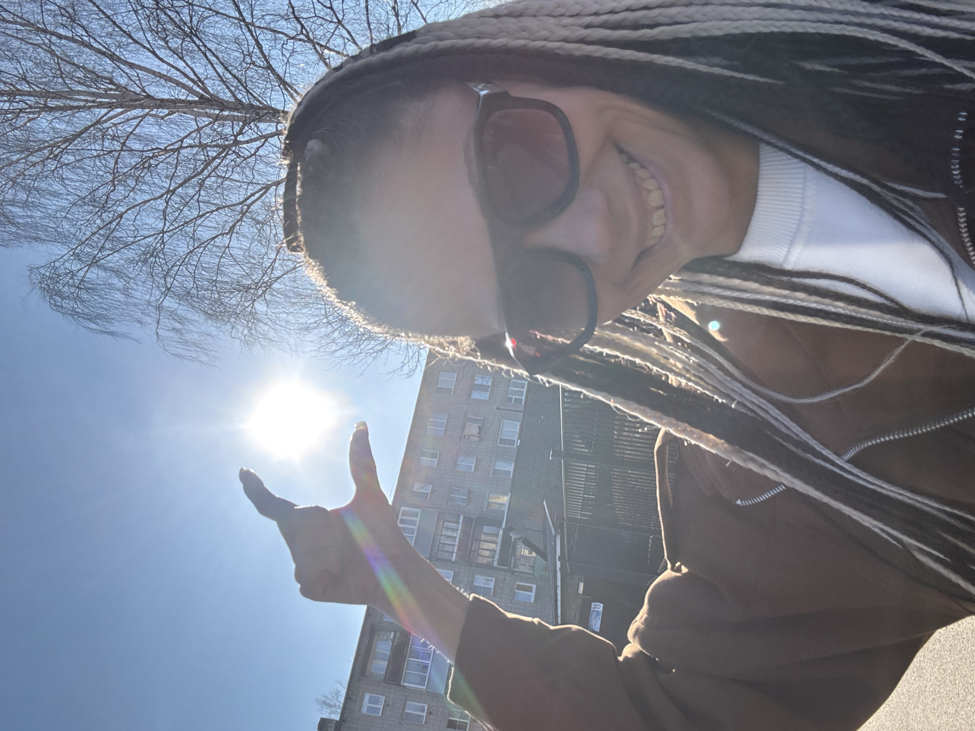 A low-angle outdoor photograph captures a person with braided hair wearing sunglasses and a brown jacket over a white top. They are giving a "thumbs up" gesture. The sun is visible in the sky, causing lens flare. A multi-story building is partially visible in the background, along with tree branches. This may be the city of Rusanovo, Russia.