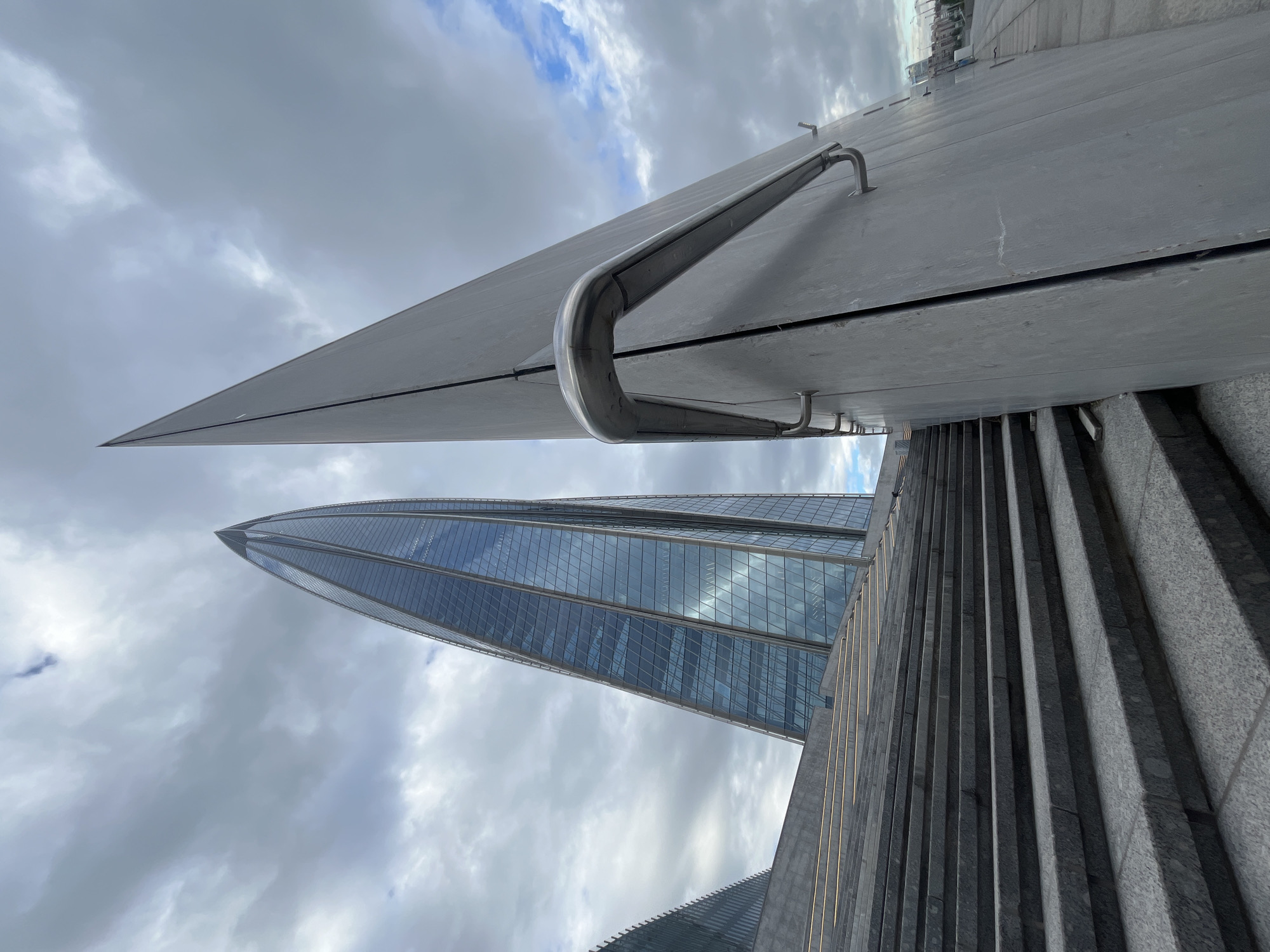 The image shows a view of the Lakhta Center, a skyscraper in Saint Petersburg, Russia, taken from an outdoor plaza. In the foreground, there are a wide set of gray granite stairs with embedded lighting running along the sides. To the right of the stairs, a large, angled, gray metallic panel is present, with a sleek silver handrail attached to it. Behind these elements, the Lakhta Center is visible, a very tall, slender, glass-faceted skyscraper that tapers to a point. Another, shorter, angular glass building is partially visible to the left of the Lakhta Center. The sky is overcast with some patches of blue sky visible, and clouds are present. The scene appears to be public space.