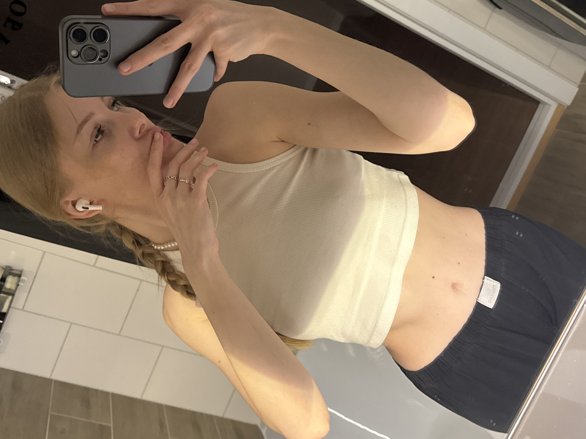 A blonde woman takes a mirror selfie in what appears to be a bathroom, given the white tiled wall and shiny surfaces, possibly a counter or tub. The woman's head is tilted to the right, and she is looking down at the camera (a dark grey smartphone with three camera lenses). Her left hand holds the phone, while her right hand is near her mouth, wearing a ring with what looks like a chain link design and another plain ring on her ring finger. She is also wearing a white earbud in her left ear and a pearl necklace. Her blonde hair is styled in a braid that extends over her left shoulder. She is wearing a light-colored, possibly white or cream-colored, ribbed crop top and dark, loose-fitting bottoms, which could be shorts or pajama pants. A white tag with unreadable text is visible on the waistband of her bottom garment. The lighting in the scene suggests an indoor setting, likely in the daytime or well-lit by artificial lights.