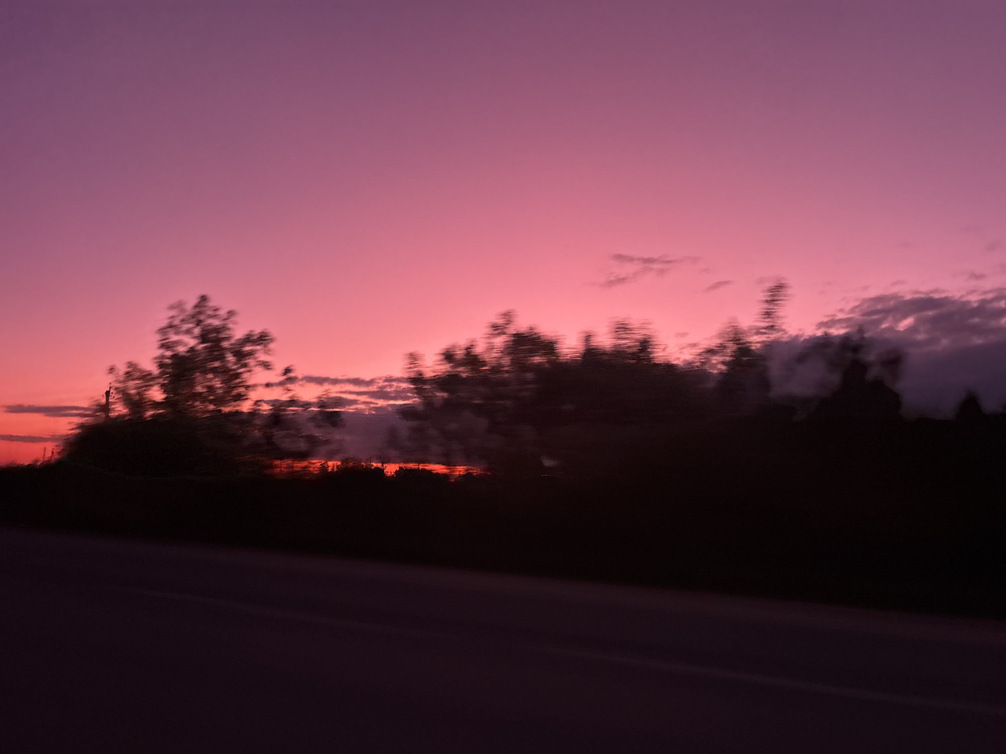 The image depicts a blurred landscape during sunset. The sky is a gradient of pink and purple. A dark silhouette of trees and foliage stretches across the middle ground. A blurred asphalt road is visible in the foreground. The image was captured with motion blur. It is hard to recognize the objects more precisely, the location is ambiguous.
