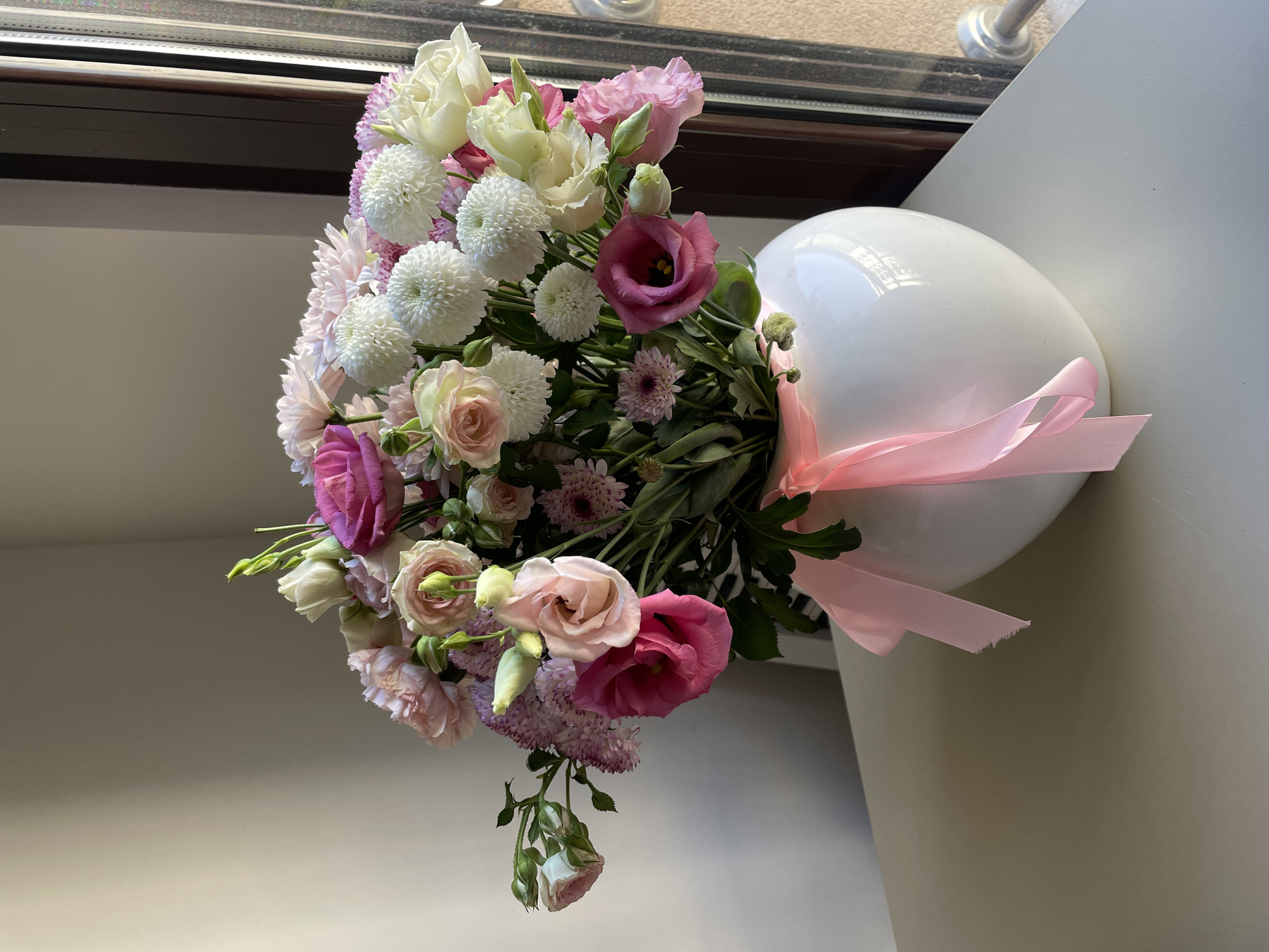 The image shows a bouquet of pink, white, and light purple flowers in a white vase with a pink ribbon. The vase is placed on a white surface next to a window with a dark brown frame. The location of the image is Boreti, Montenegro.