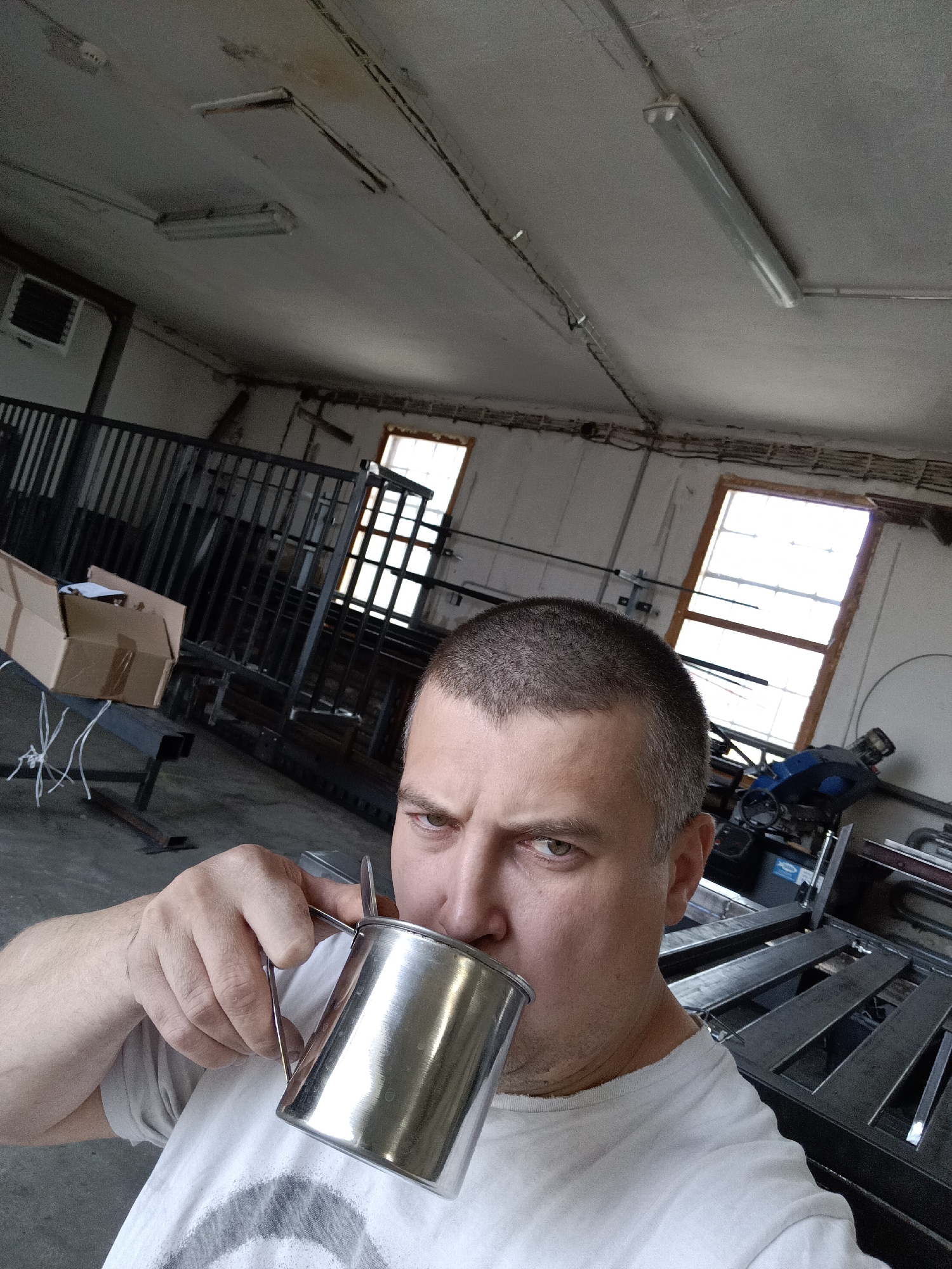 In the city of Dalovy, Czechia, a man with a buzz cut is captured in a selfie, holding a metallic mug to his lips as he drinks. He wears a white t-shirt and is positioned in what appears to be a workshop or industrial space. The background reveals metal structures, including a railing and what seems to be a workbench or fabrication table. Two windows with grid-like panes are visible, admitting natural light. Overhead, fluorescent lights illuminate the ceiling, and an air conditioning unit is mounted on the wall. A cardboard box rests on a metal beam in the left portion of the frame.