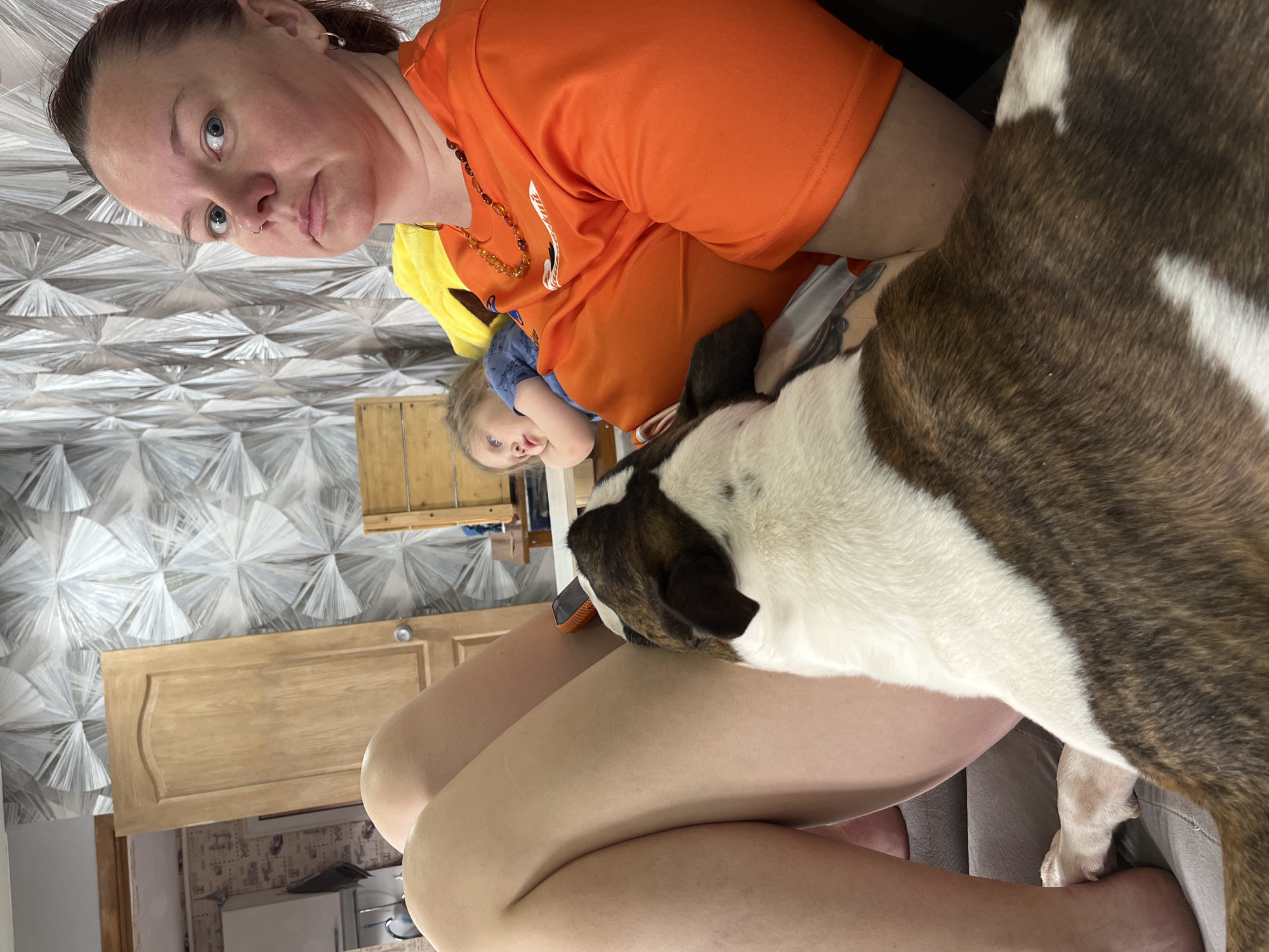 The image, which is rotated 90 degrees counter-clockwise, captures an indoor scene in Chaikovsky, Russia, featuring a woman, a child, and a dog.
In the upper-left portion of the frame, an adult woman with light skin, dark hair pulled back, and light blue eyes looks directly at the camera. She wears an orange short-sleeved top, an amber beaded necklace, and has a nose piercing. Her expression is calm and neutral.
Below her head, towards the center-left, a young child is partially visible, wearing a blue top with a yellow layer underneath. The child also looks towards the camera with a neutral expression. On the right side of the frame, a brindle and white dog, possibly a Bull Terrier mix, rests comfortably against the woman's legs, with its head turned away from the camera. An orange tag is visible on what appears to be its collar.
The background, visible behind and above the subjects, features a distinctive grey and white geometric patterned wallpaper. A light-colored wooden cabinet or counter is also seen below the patterned wall. The overall impression is a relaxed, everyday moment during daytime in a home setting. No visible text is present in the image.