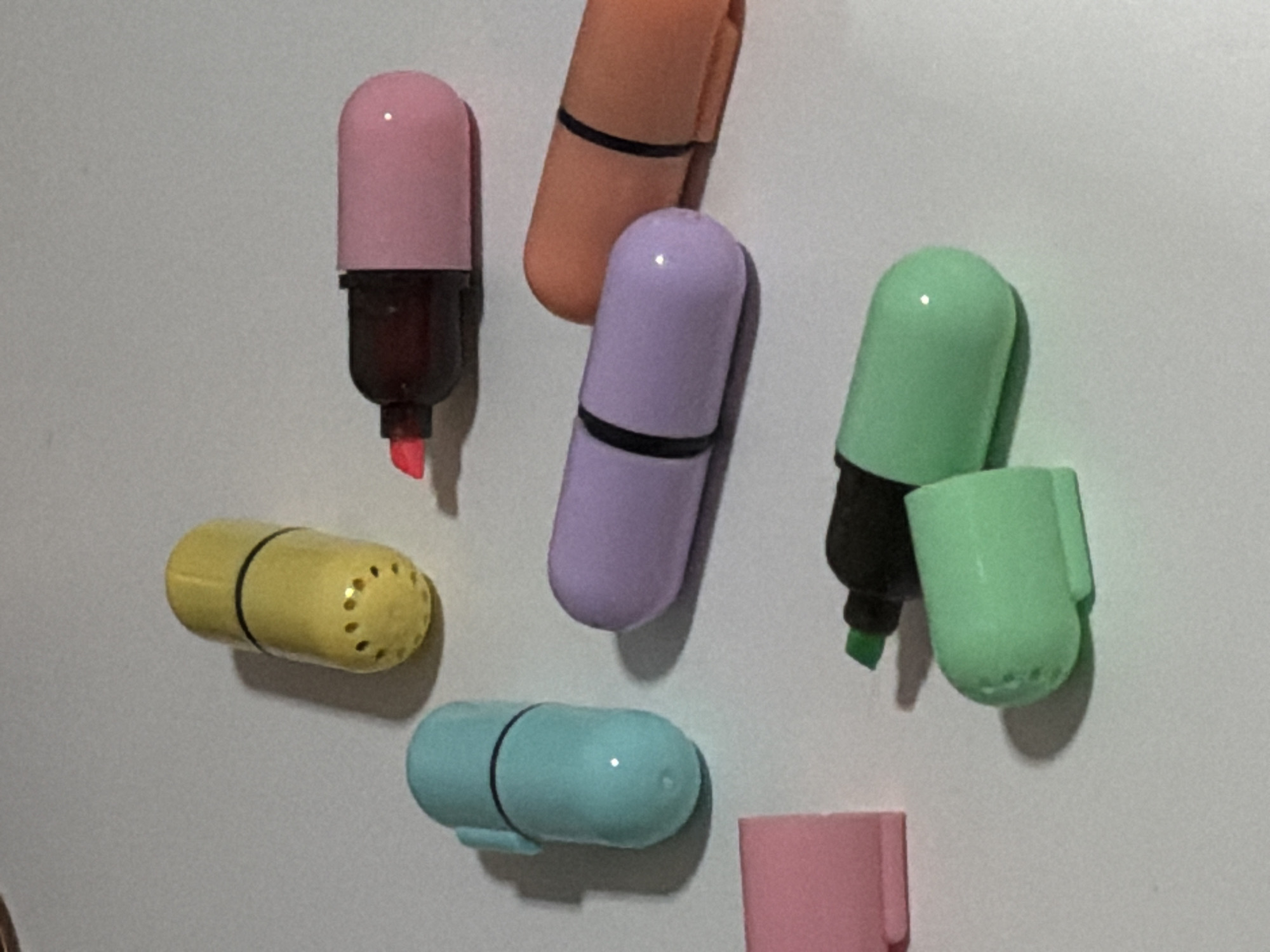 The image shows a collection of colorful capsule-shaped highlighters scattered on a white surface. There are highlighters in yellow, light blue, pink, purple, orange, and green. Some of the highlighters are closed, while others are open, revealing their colored tips. The yellow highlighter has a series of small dots on its cap. The pink highlighter has a red tip. The green highlighter is shown with its cap removed, revealing a green tip. The image does not provide any information about the location of this scene in Moscow, Russia, or any details about people, actions, or interactions. The environment appears to be a plain, indoor surface, likely a desk or table.