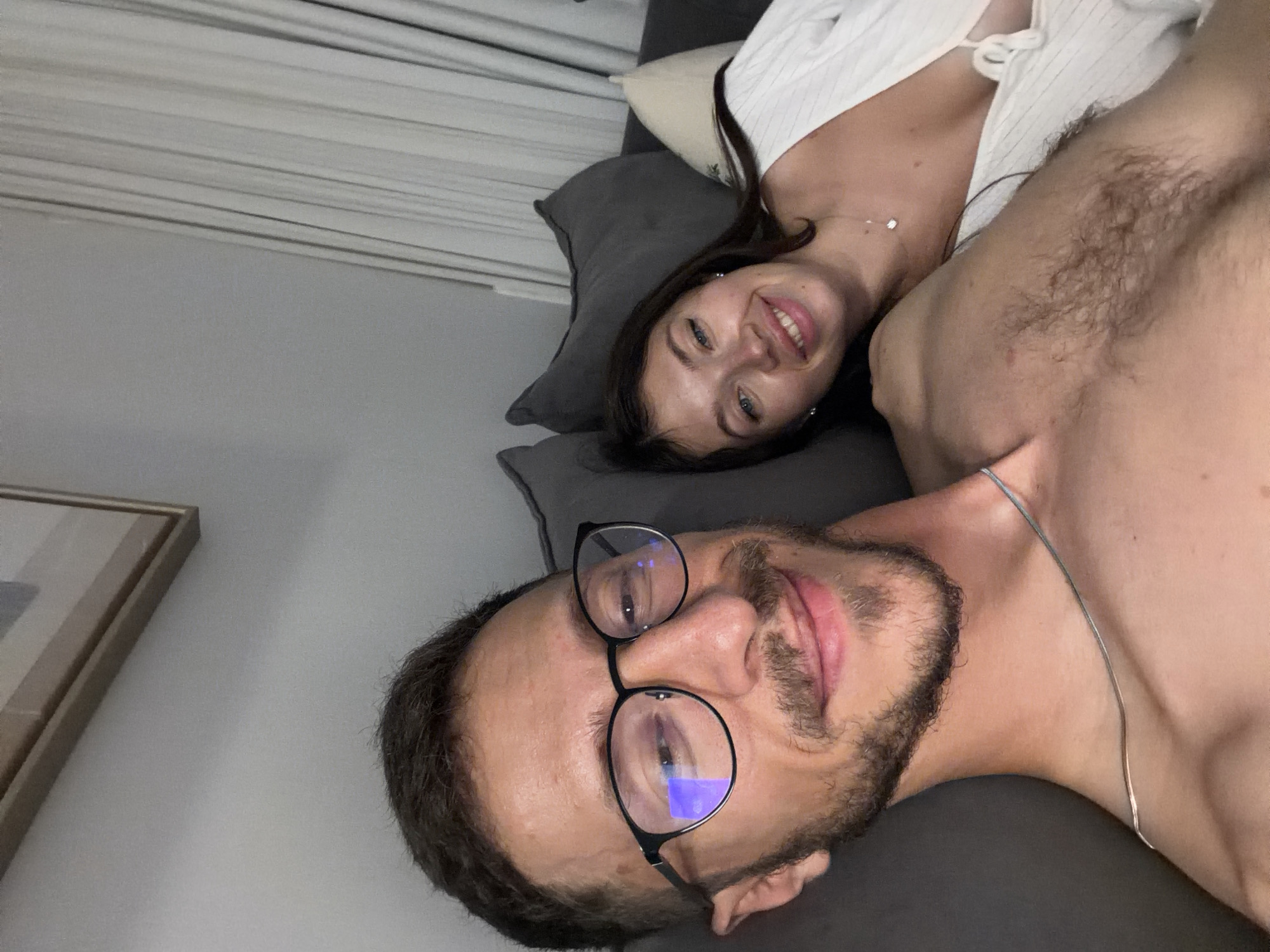 This is a selfie of a man and a woman on a couch. The man is in the foreground, wearing glasses and a thin necklace. He has a beard and is smiling. The woman is behind him, leaning on a pillow, and smiling. She has dark hair and is wearing a white top. The couch is dark gray, and there are light-colored curtains in the background. The scene appears to be indoors, in a residential setting. There is no discernible information to suggest the location is the city of Tel-Aviv, Israel.