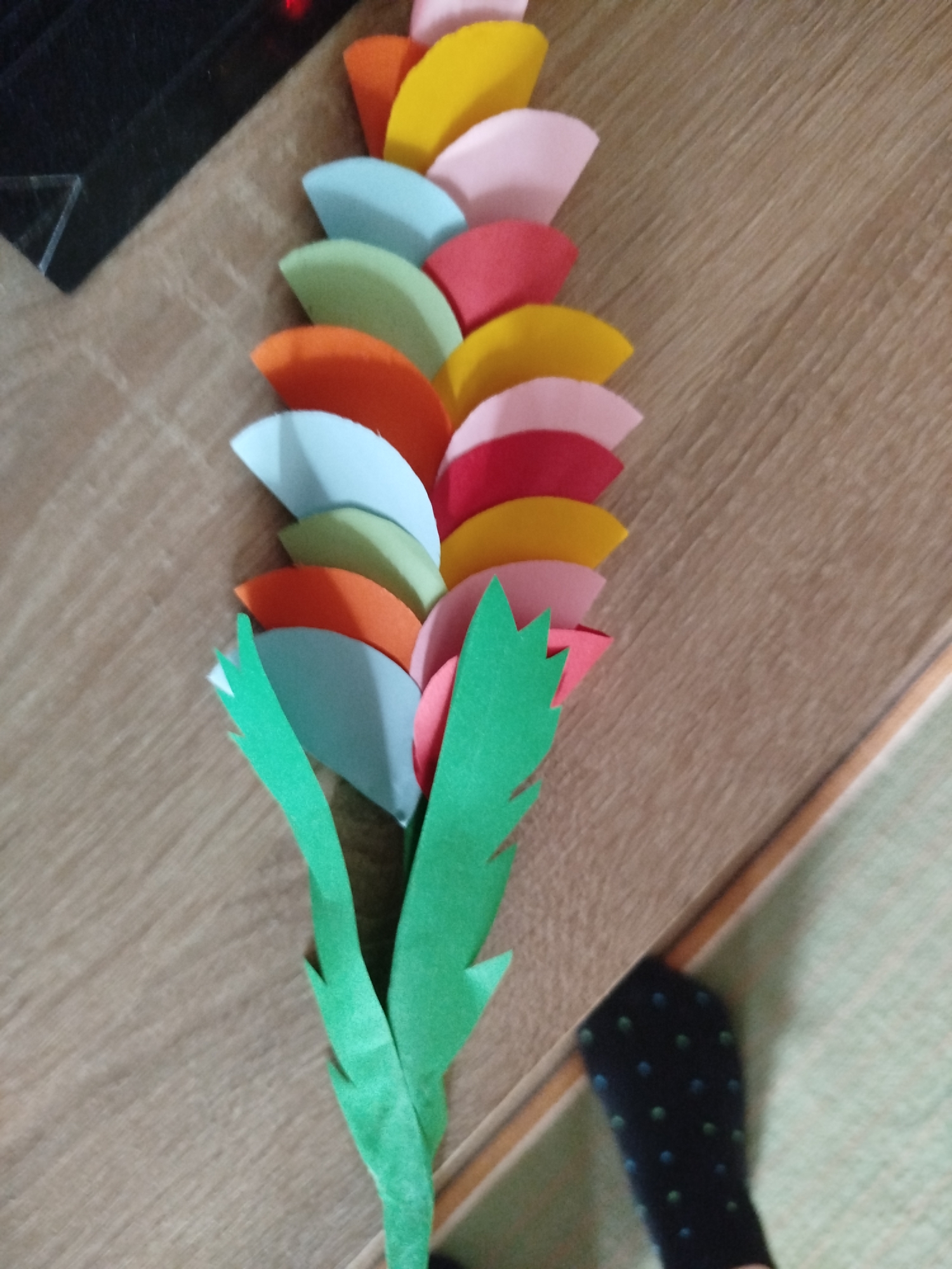 The image displays a close-up, high-angle shot of a colorful paper craft resembling a flower or a spray, laid out on a wooden surface. The craft consists of multiple layered, semicircular cutouts in various shades of yellow, pink, red, orange, and light blue, arranged in a staggered, fan-like pattern. Two green paper leaf-like shapes are attached to the bottom of the craft, one larger and more prominent than the other. The wooden surface appears to be a table or floor with a prominent wood grain pattern. In the lower right corner of the image, a black sock with a pattern of small green and blue dots is visible, suggesting the presence of a person. The background shows a dark, possibly black object in the upper left corner, with a triangular reflective surface. There are no visible landmarks or specific environmental features that would indicate a location within Kryvaselski Sel'ski Savet, Belarus.