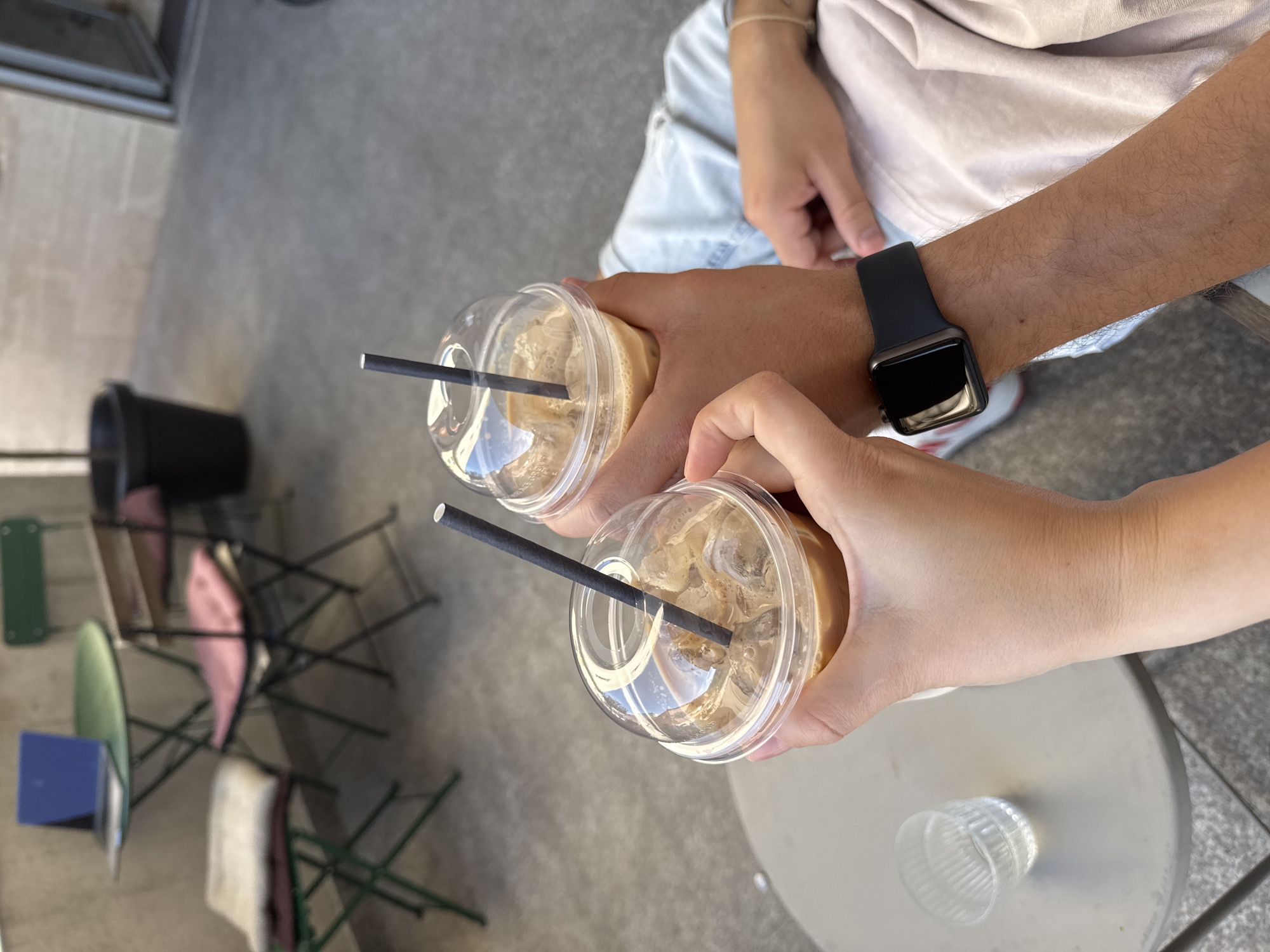 The image depicts two individuals clinking clear plastic cups of iced coffee in an outdoor setting in Budva, Montenegro. A lighter-skinned hand, presumably female, holds the left cup, while a darker-skinned, hairy forearm and hand, wearing a black smartwatch, holds the right cup. Both cups contain a light brown beverage with ice cubes and are topped with clear domed lids and dark paper straws. The individual on the right is partially visible, wearing a light blue denim shirt and light-colored trousers. In the blurred background, an outdoor cafe area is present, featuring a circular green metal bistro table with an open blue laptop. Various folding chairs are arranged around the table, including a green metal chair with layered white and maroon cushions in the mid-ground, and another dark metal chair with a pink cushion. A large black planter is positioned in the right background. A small, clear, ribbed glass rests on a light-colored, round table, partially visible in the bottom left foreground. The ground is a textured grey surface, and a light-colored, textured wall forms the distant background.