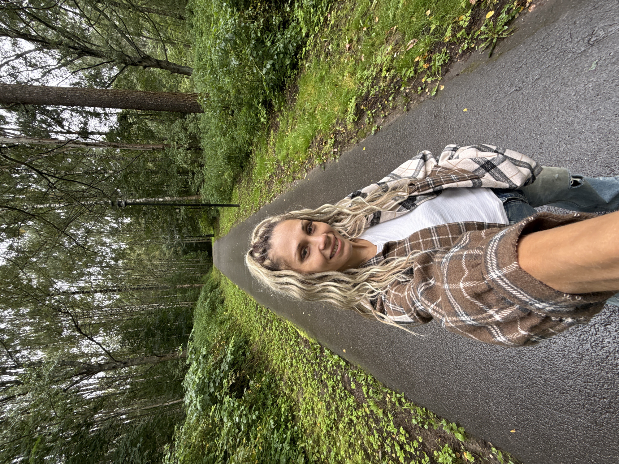 A blonde woman with wavy hair is taking a selfie on a wet asphalt path in a park in Dumino, Russia. She is wearing a white tank top and a brown and white plaid jacket. She has on ripped blue jeans. The path is lined with trees and green foliage. The sky is overcast.