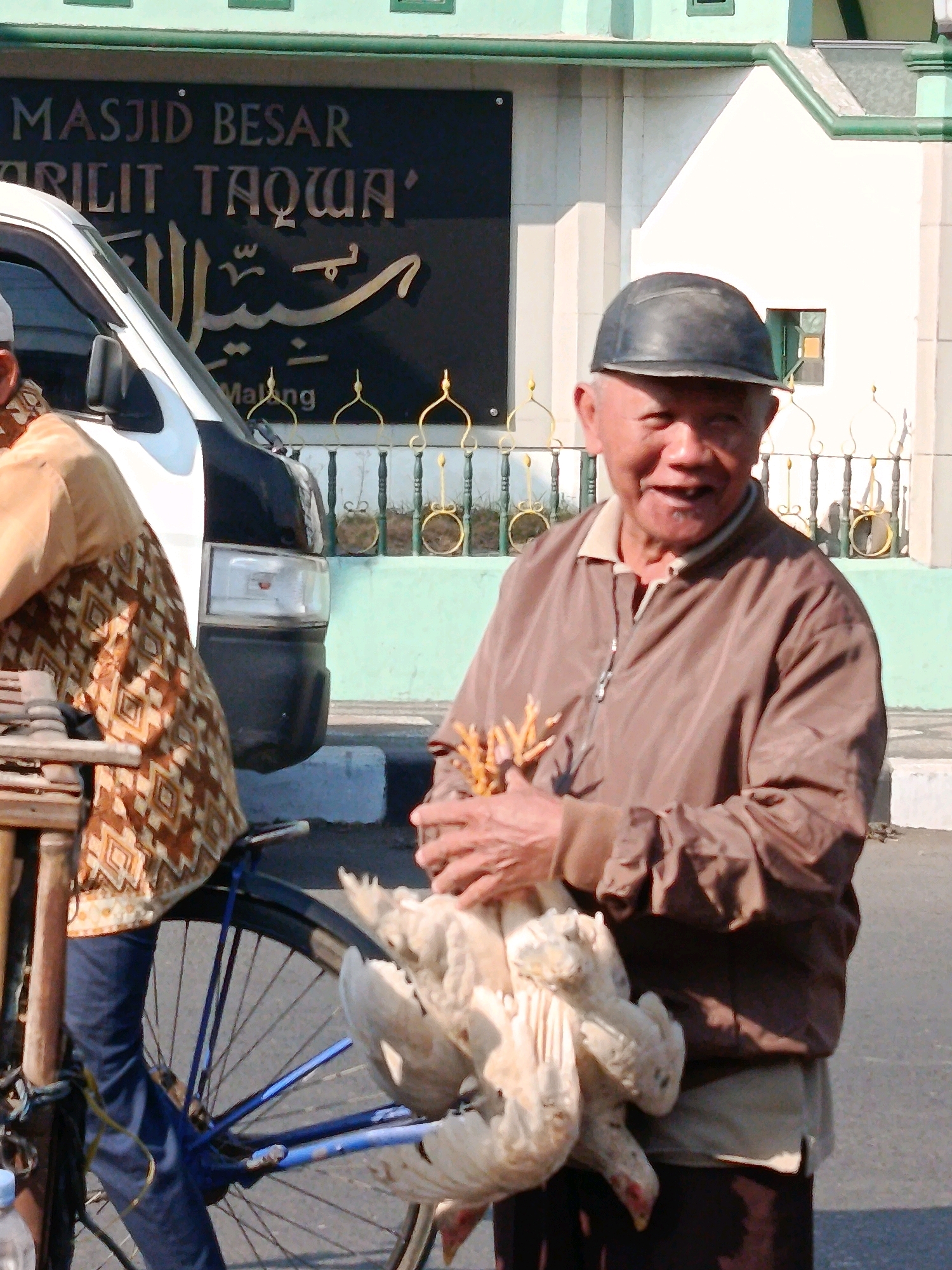 An outdoor, eye-level, medium shot captures a smiling, elderly Asian man wearing a dark cap and a brown zippered jacket, holding two dead chickens with light-colored feathers. The man stands in front of a white and green building with ornate railings. To his left, partially visible, is the side of a white van with a black stripe. Behind the man, a sign reads "MASJID BESAR" and "ARILIT TAQWA" in gold lettering on a black background, with Arabic calligraphy above. The man's hands are clasped around the chickens, which are held upside down with their feet pointing upwards. He appears to be in a commercial or public area, possibly a market, given the presence of the van and the transaction implied by the chickens. The lighting suggests it is daytime, with shadows indicating the sun is to the right. The overall scene is consistent with a street vendor in a town like Bululawang, Indonesia.