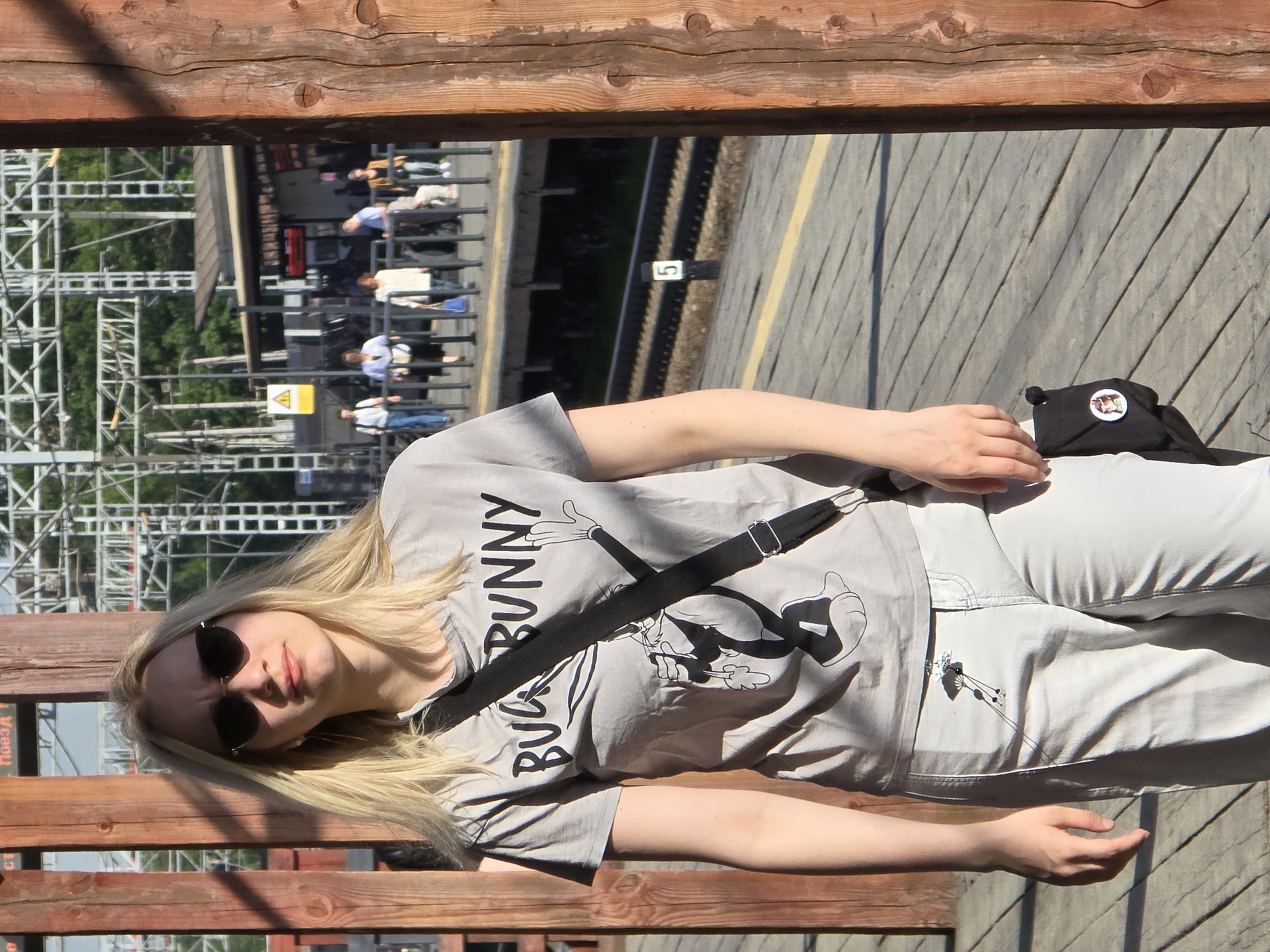 A person with blond hair and sunglasses is resting on a wooden bench at what appears to be a train station. The individual is wearing a gray T-shirt with a Bugs Bunny design and light-colored pants. A black bag is next to them on the bench. A railway platform and tracks are visible in the background, with people walking along the platform. A sign with the number "5" is also visible. The image was taken at a location in the city of Khimki, Russia.
