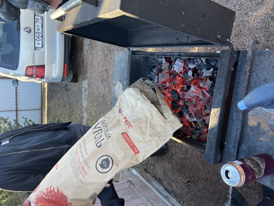 The image depicts an outdoor scene in Заозерное Снт, Russia, centered on a black metal brazier containing glowing red and gray charcoal. A person, identified by a partially visible gloved hand and arm, is pouring birch charcoal from a large brown paper bag labeled "БЕРЕЗОВЫЙ УГОЛЬ" (Birch Charcoal) by "Шефрепс HORECA LINE" into the brazier. An open aluminum beverage can with Cyrillic text "ПИДС" is positioned on the left edge of the brazier's frame, adjacent to a blue and gray handheld torch on the right. In the background, a silver Volkswagen Touran minivan, bearing Russian license plate "У 068 МТ 98 RUS", is parked. Portions of two additional individuals are discernible: one standing to the left of the brazier, and another near the vehicle. The ground consists of dry soil with minimal vegetation, and a light-colored corrugated metal fence is visible behind the vehicle.