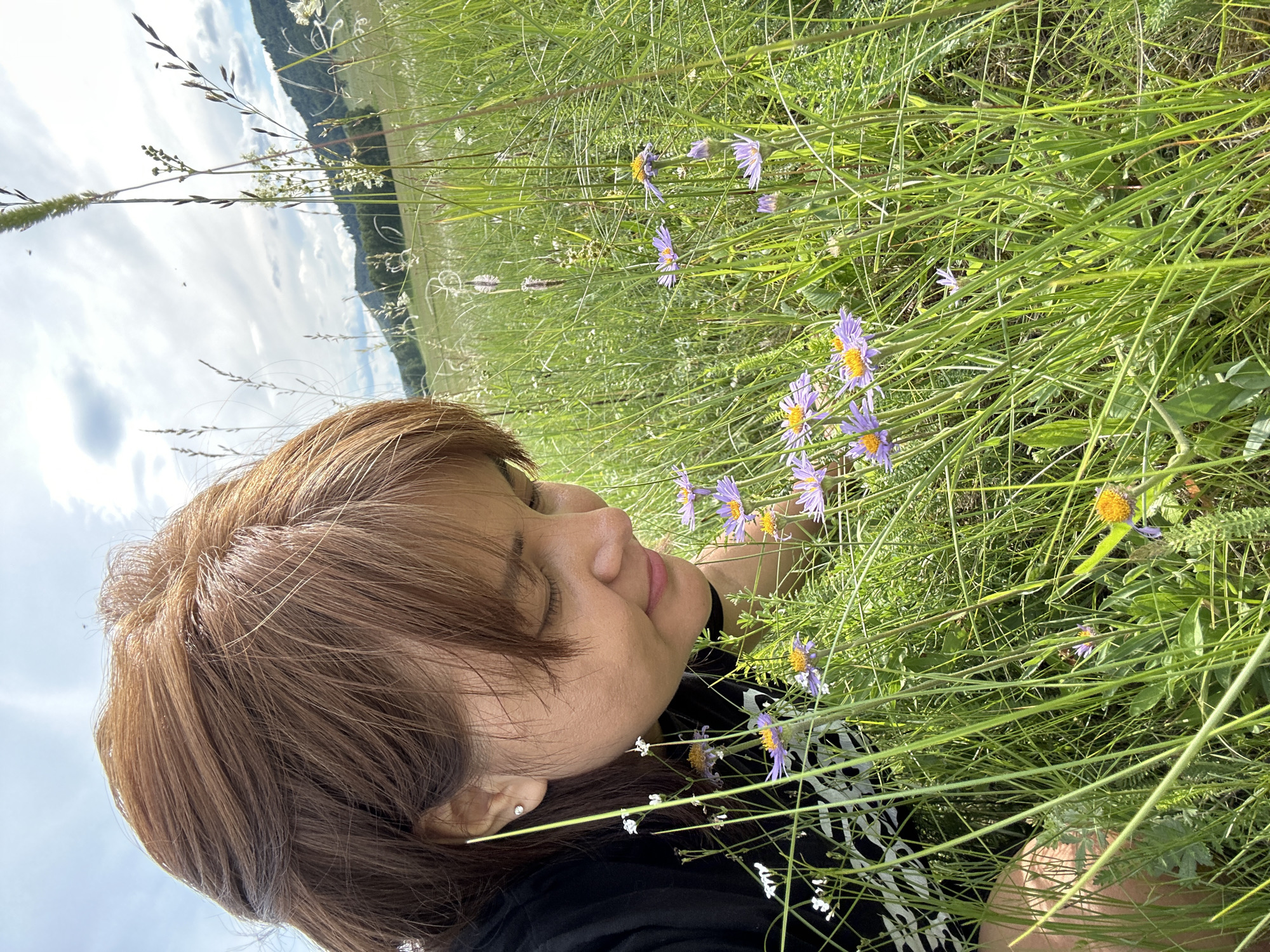 This is a media file of a person lying in a field of tall green grass and wildflowers. The person, with brown hair and wearing a black top, is lying on their side with their eyes closed, appearing to be relaxing or sleeping. Purple and yellow wildflowers are scattered throughout the grass, some in focus and others blurred by the depth of field. The background shows a slightly hazy landscape with distant hills and a cloudy sky. The lighting suggests it is daytime, likely during the day. There is no visible text in the media file.