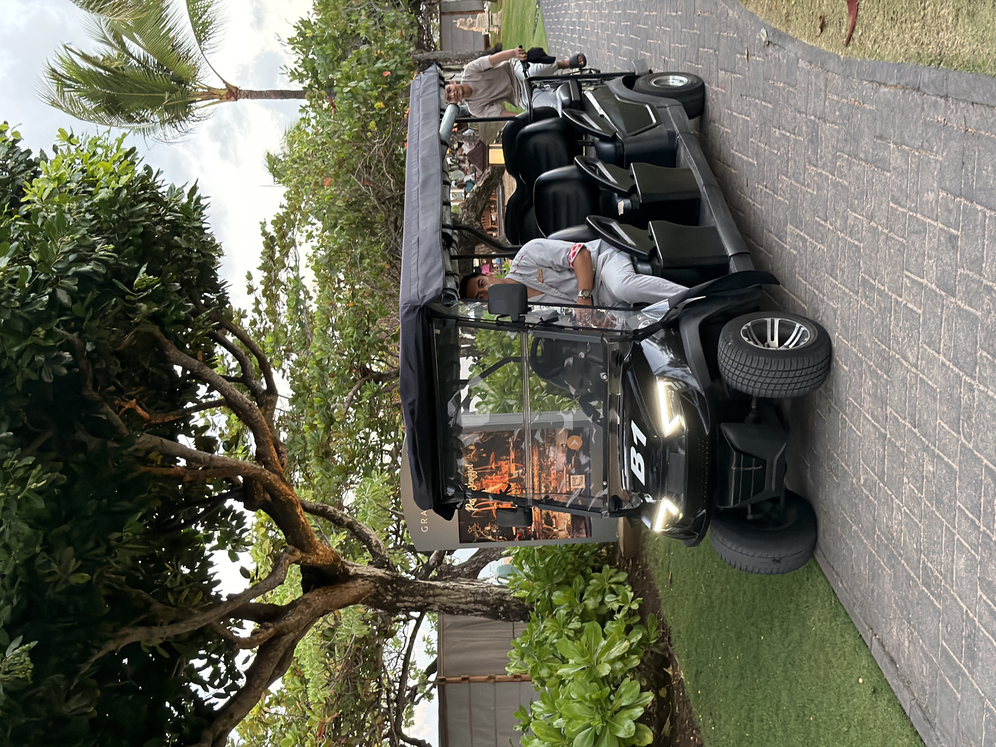 A black golf cart with a grey canopy is parked on a paved walkway next to a grassy area. There are three people in the golf cart. The person in the front passenger seat is wearing a light-colored polo shirt and grey pants. The person in the driver's seat is wearing a light-colored shirt and black shorts. A third person, wearing a light-colored shirt, is seated behind them. The golf cart has the number "B1" visible on the front.
In the background, there are trees with green foliage and a glimpse of a building with thatched roofing. The scene appears to be outdoors, likely in a tropical setting, given the presence of palm trees and lush vegetation. The lighting suggests it is daytime, possibly late afternoon or early evening. The overall atmosphere appears relaxed and leisurely. The paved walkway is made of interlocking gray bricks.