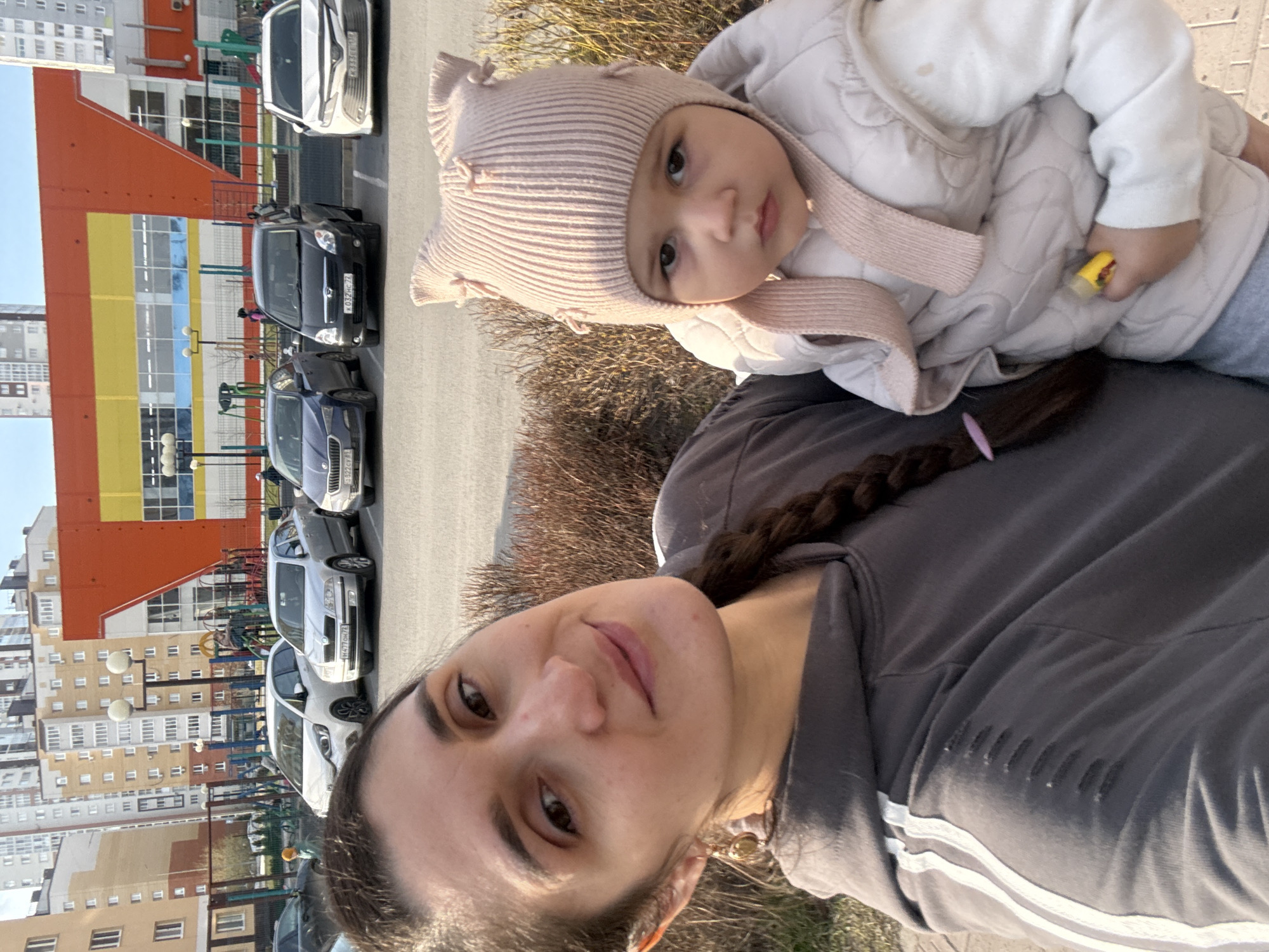 The image presents a selfie-style portrait of a woman holding a baby outdoors during a bright, sunny day. The woman, in the foreground, has dark hair styled in a braid with a pink clip and wears a dark grey top with white stripes on the sleeve. She looks towards the camera with a gentle expression.
The baby, held in the woman's arms, wears a light pink knitted hat with decorative ties and a light-colored padded jacket, suggesting cool weather. The baby also looks towards the camera with a neutral, curious gaze.
In the background, a row of parked cars is visible on a paved area, likely a street or parking lot. Several cars display Russian license plates. Notably, multiple cars have the "72" region code (e.g., "K032HC 72", "322CY 72", "H470AO 72"), which corresponds to the Tyumen region, aligning with the Tobolsk location context. One car has a "154" (Novosibirsk region) license plate.
Beyond the vehicles, multi-story residential buildings line the horizon, with one prominent structure featuring vibrant orange and yellow sections. Colorful playground equipment can be seen between the cars and buildings. Sparse, dry bushes are visible along the edge of the paved surface. The scene suggests a casual outing in a residential area.