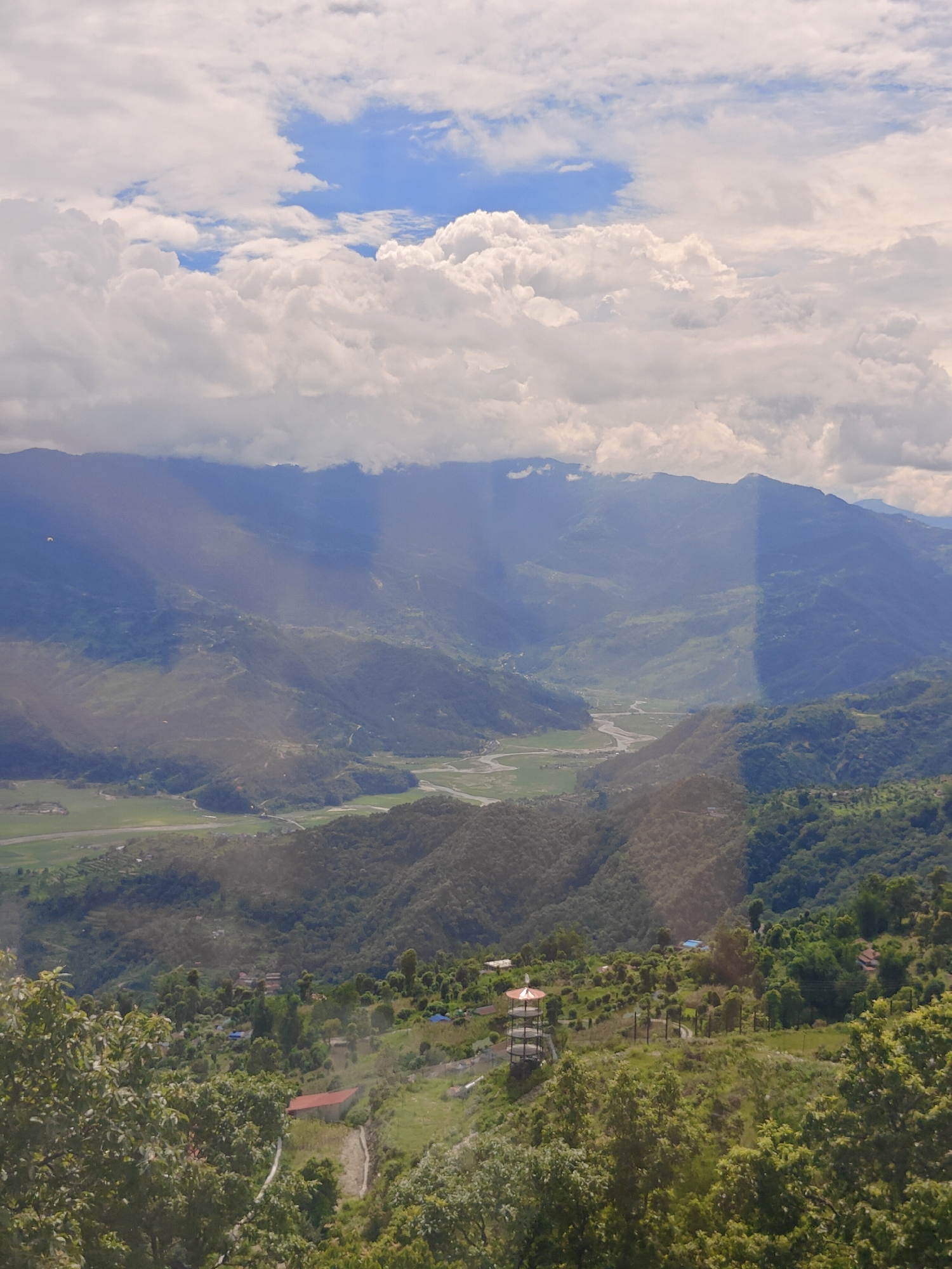 The image is a high-angle, wide shot of a mountainous landscape, likely in Pokhara, Nepal, under a partly cloudy sky. In the foreground, dense green foliage and trees frame the bottom of the image. A small, red-roofed building is visible in the mid-foreground, nestled among the trees. Further back, on a hillside, is a distinctive multi-tiered tower structure with a red roof, surrounded by a fence. A winding path leads towards it. Scattered across the mid-ground are small clusters of buildings, some with blue roofs, interspersed with green fields and more trees.
The middle ground transitions into a lush, undulating valley with a river or stream winding through it. The valley is surrounded by steep, green hillsides that rise to meet imposing mountains in the background. Sunlight pierces through gaps in the clouds, creating dramatic shafts of light that illuminate patches of the mountains and valley, casting shadows. Two small, white paragliders are visible in the sky above the mountains. The overall impression is one of a vast, natural landscape dotted with human settlements and recreational activities.