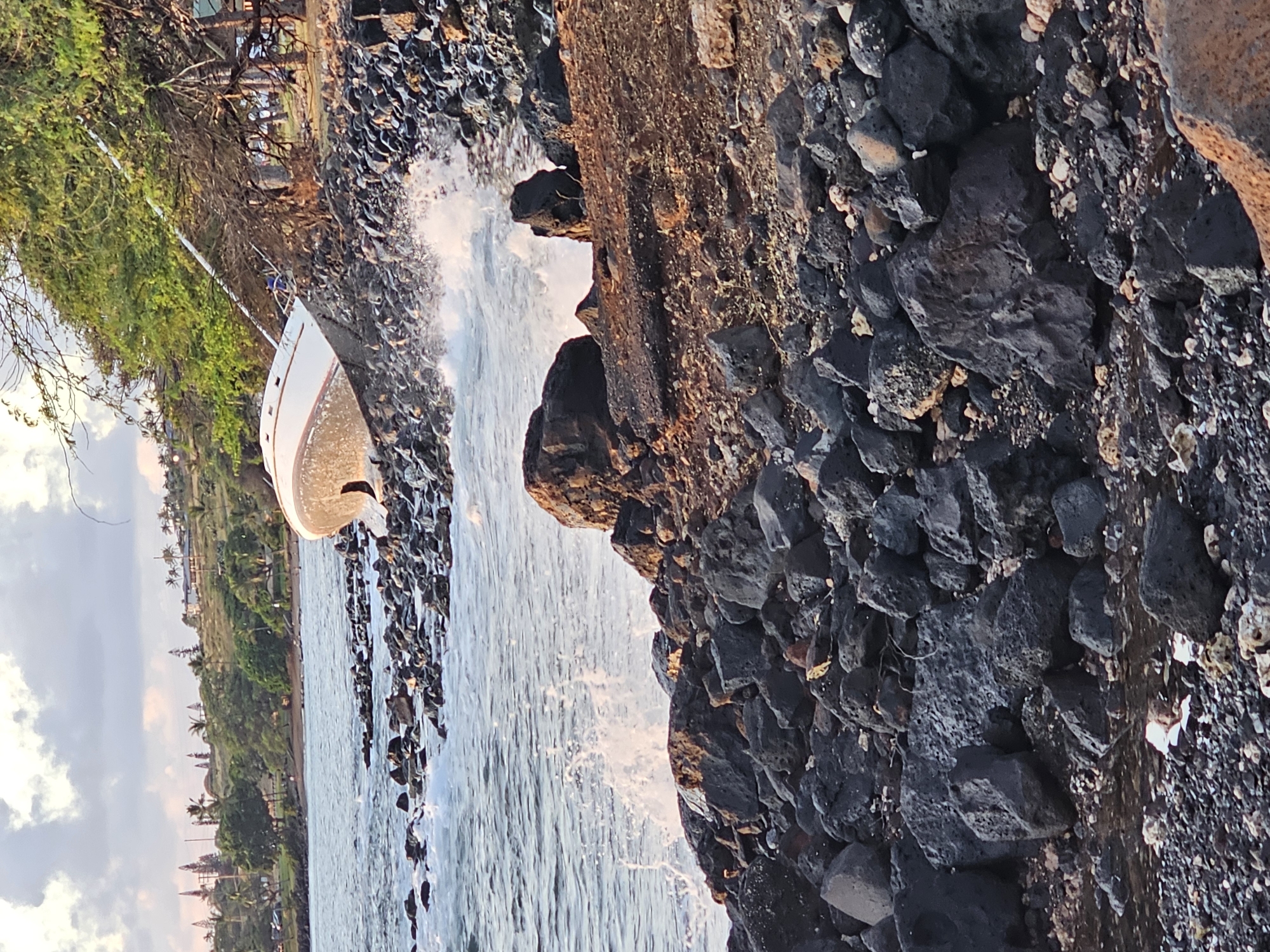 A white and red sailboat is beached on a rocky shore near Lahaina, Maui, Hawaii. The boat is tilted on its side, with its hull resting against a concrete structure and rocks. Waves are crashing against the shore, creating spray. The background shows a tree-lined coastline with hills and buildings. The sky is overcast with some visible clouds.