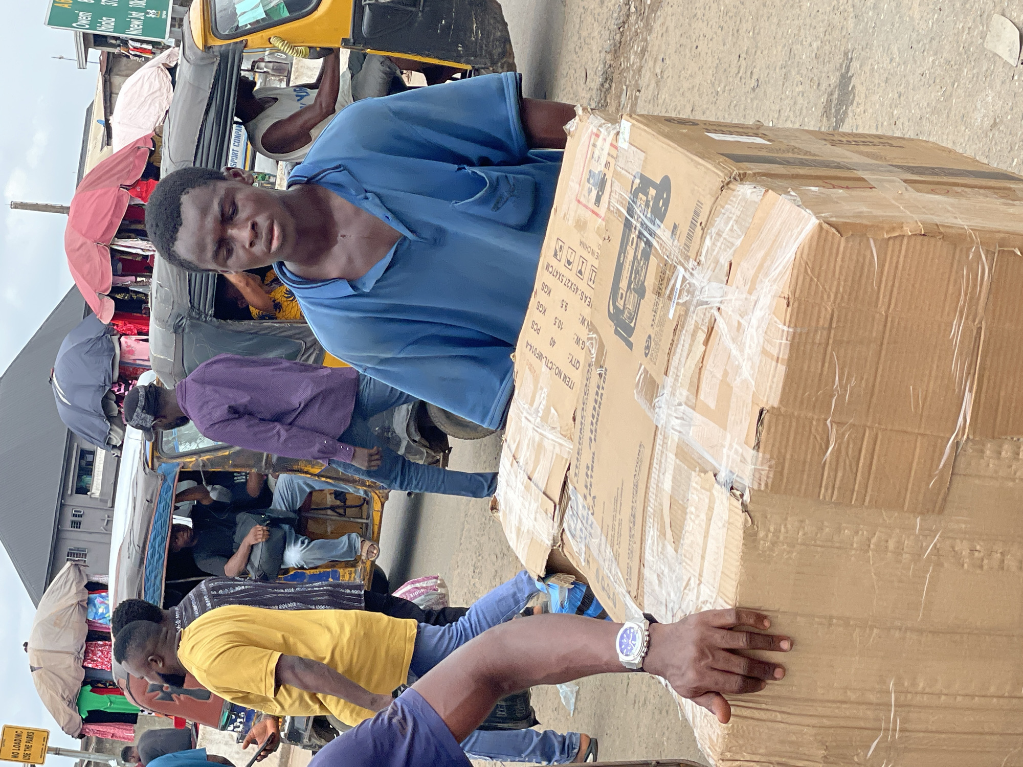 A man in a blue shirt carries a large cardboard box in Onitsha, Nigeria. A yellow tricycle is behind him, filled with passengers. Other pedestrians walk nearby. A sign reads "Oweni... Nnewi Jnt 10kc." Another sign prohibits loading in the park area.