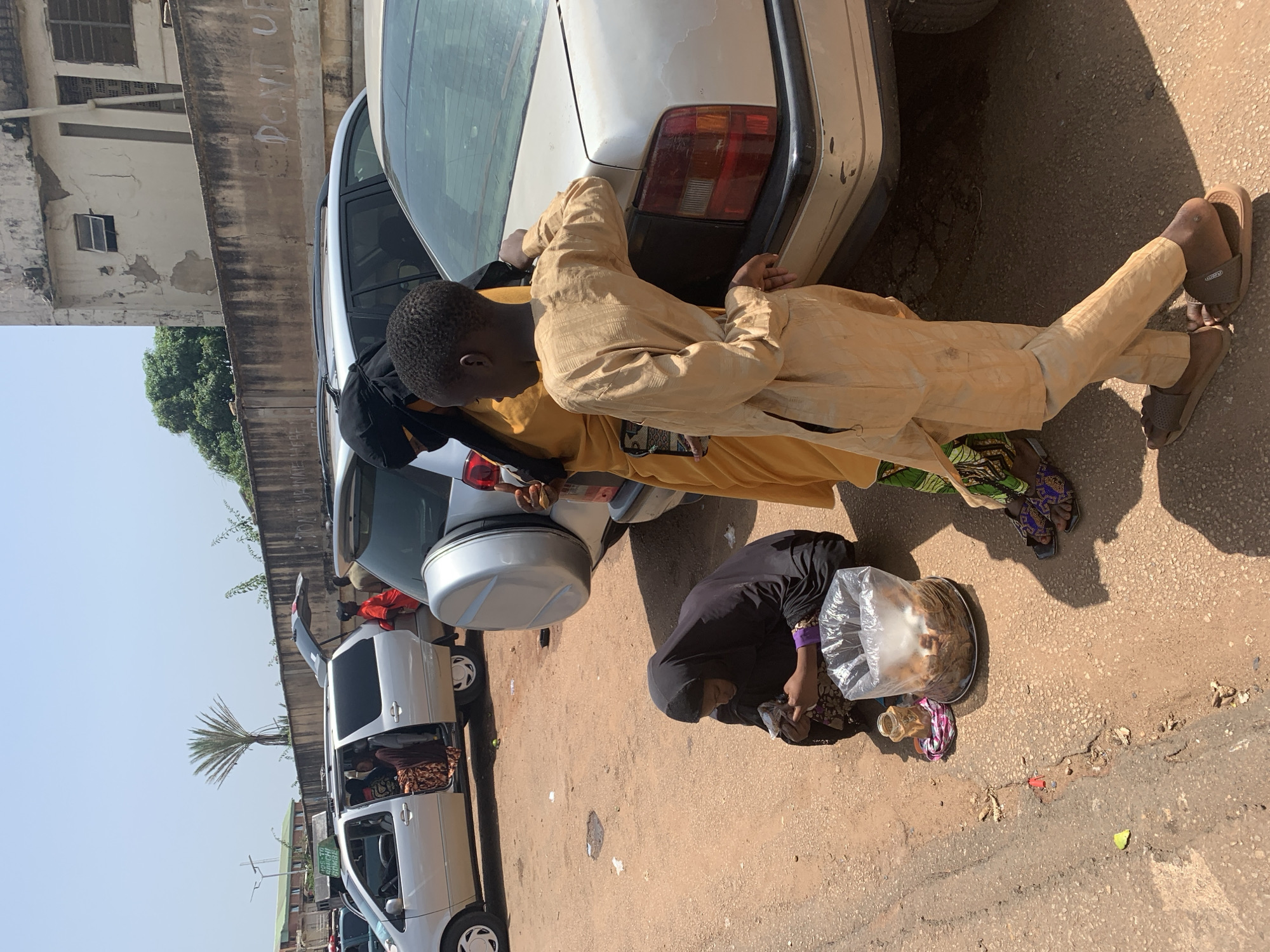 The image depicts a street scene in Jos, Nigeria, under a clear, sunny sky, suggesting it's daytime. In the foreground, a person dressed in a light brown, traditional outfit leans against a white car, with their legs crossed. They are wearing dark sandals. To the right, a person wearing a black hijab and a patterned skirt is crouched down, tending to a plate of food covered with plastic wrap, and a small bottle. The food appears to be bread or pastries. Beside them, a small, colorful fabric is laid out.
In the background, a white car is parked next to a concrete structure. Another white van with its doors open is visible to the left, with people inside. Further in the distance, a palm tree stands tall against the sky. The ground is dusty and uneven, typical of a street in this region. The overall impression is one of everyday life and commerce taking place outdoors. There is some graffiti visible on the concrete wall in the background.
