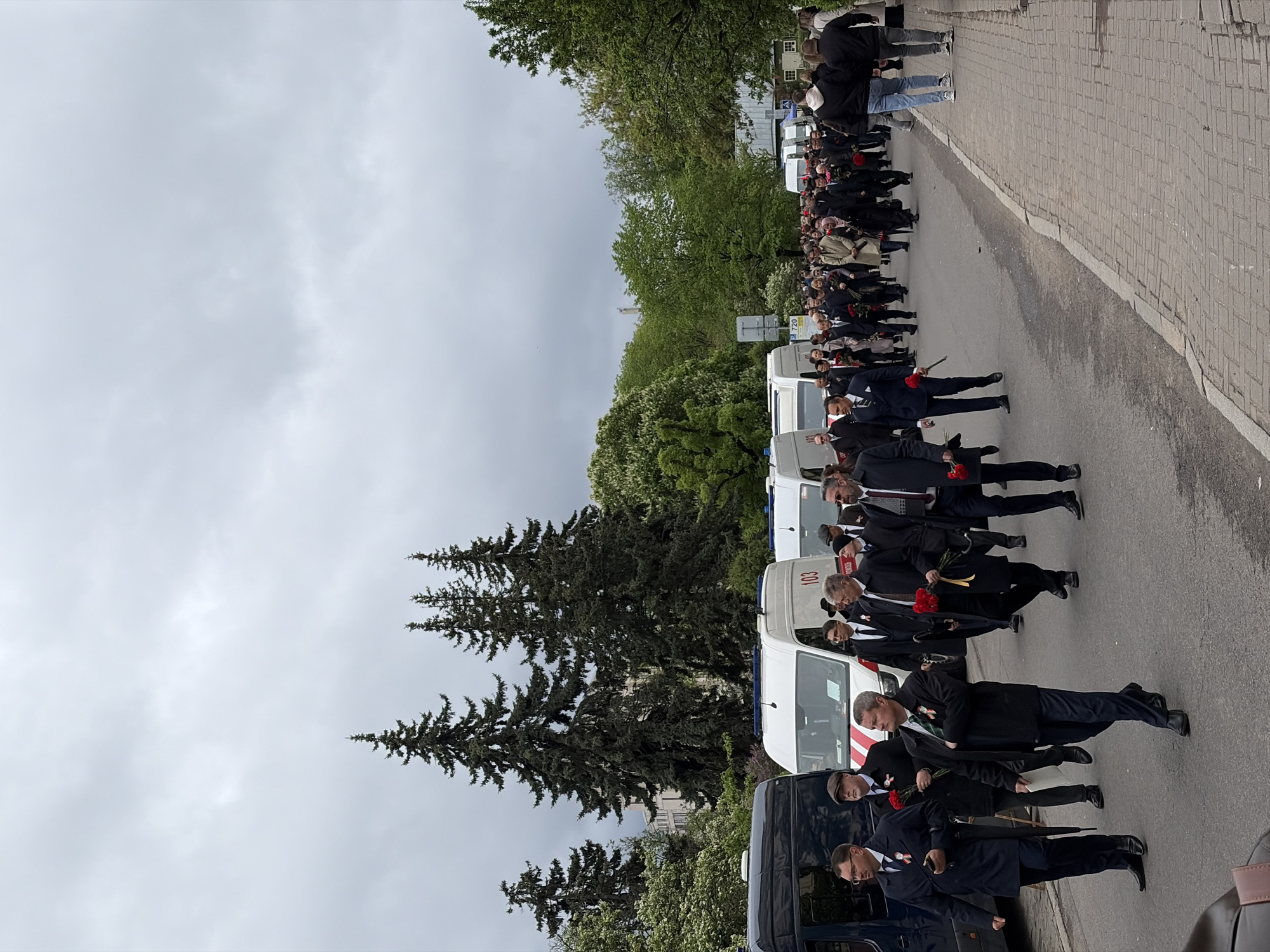 A long line of people dressed in dark suits and holding red flowers walks along a street. Several vans, including one with the number "103" visible, are parked alongside the street. A group of people stand on a paved area at the far end of the line. Tall trees line the street, and the sky is overcast. The location is in the city of Unknown, Unknown.