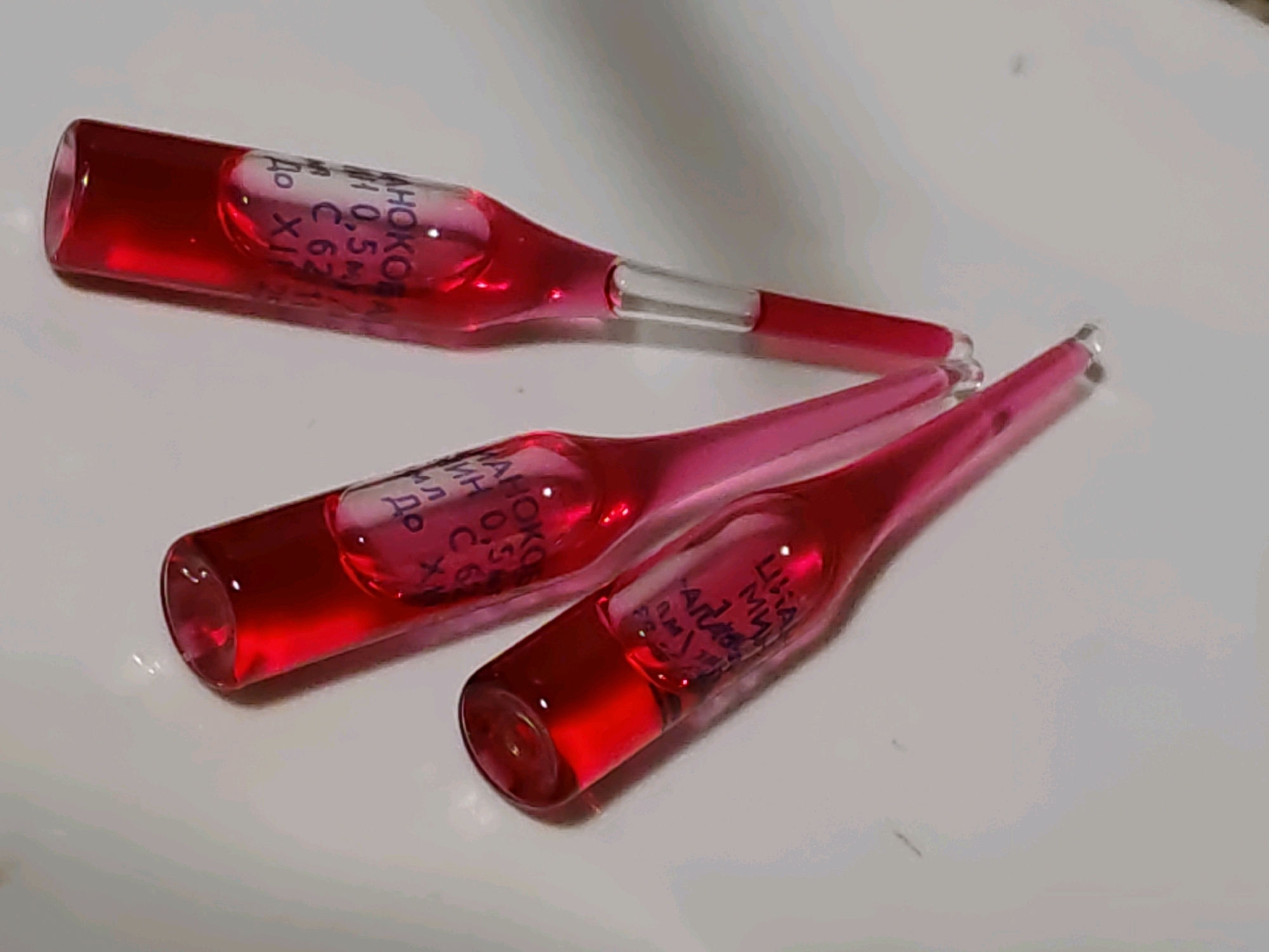 Three red-colored glass ampoules containing a red liquid are lying on a white surface. The ampoules are of varying sizes and are arranged in a diagonal line from the top left to the bottom right of the image. Text is visible on two of the ampoules, but it is difficult to read due to the image resolution and angle. The white surface appears to be a flat, non-reflective material, possibly a countertop or table. The lighting is soft, casting subtle shadows from the ampoules. There are no people or any indication of a specific location within the city of Rostov-on-Don, Russia, in this image.