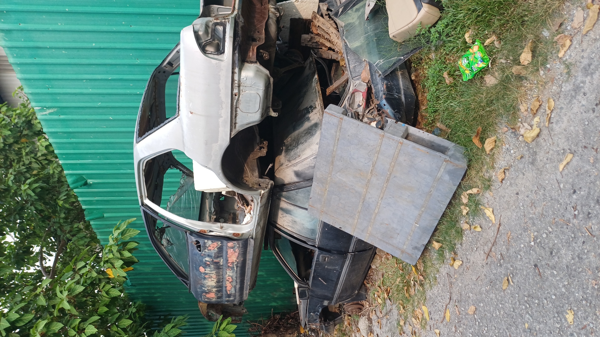 In Kuala Lumpur, Malaysia, a scene of disused vehicles is depicted. Two car bodies are stacked atop one another, positioned in front of a green corrugated metal fence. The upper car, predominantly silver, is missing its hood, doors, and roof, revealing its skeletal interior. Its lower section displays signs of rust and wear, particularly on the driver's side door panel, which is a peeling black. Below this, a black car, also stripped of many components, lies on its side. Its doors and windows are present, though its front end appears significantly damaged. A large, flat metal sheet rests diagonally across the lower car and the ground, obscuring some of the view. The ground is a mixture of dirt, gravel, and fallen leaves, with patches of green grass. A small, colorful wrapper lies on the grass to the right of the metal sheet, near a cream-colored car seat. Trees with green foliage frame the top left of the image, with branches extending towards the fence. The overall impression is one of abandonment and decay, likely within an impound lot or a junkyard.