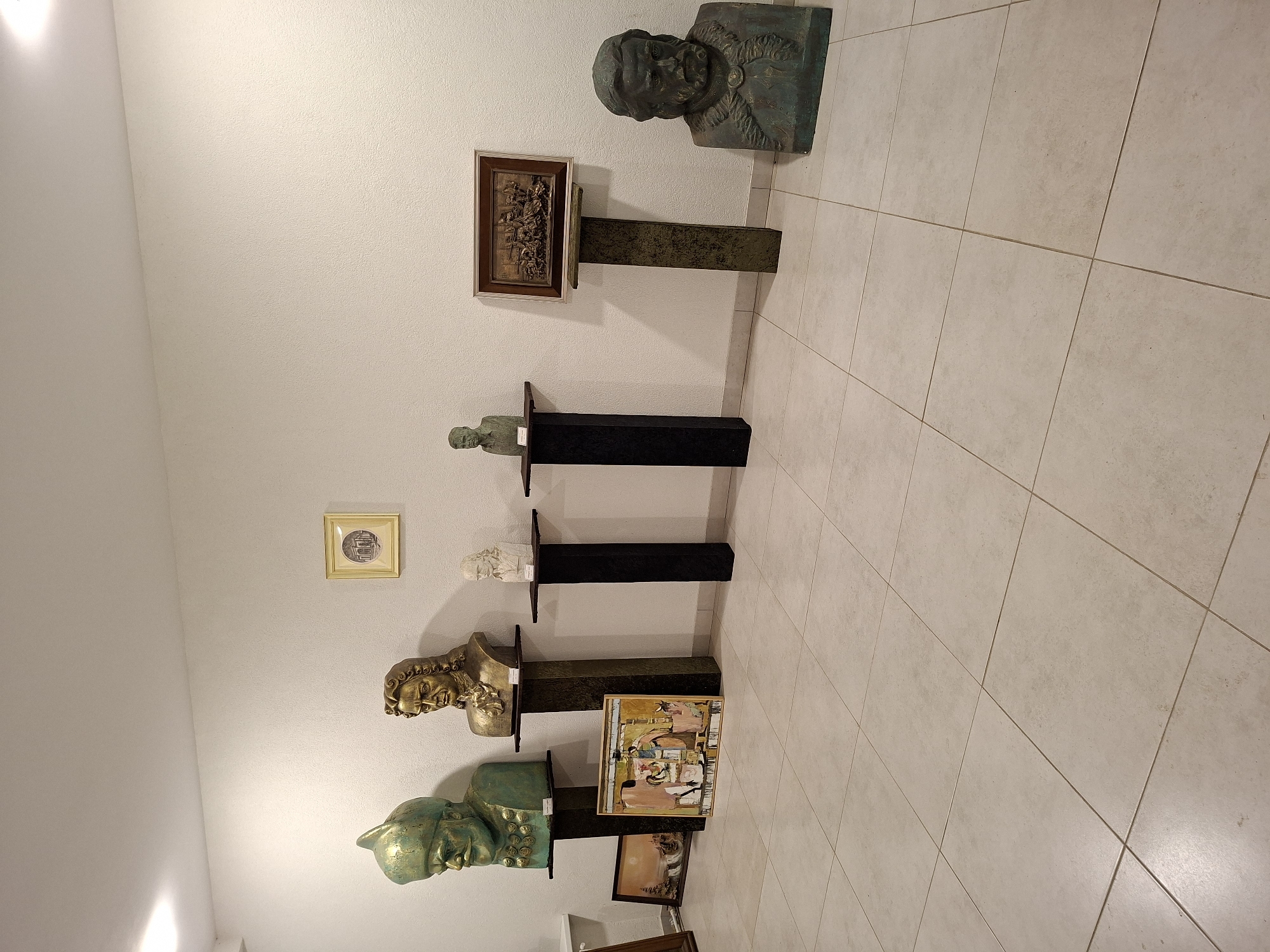 The image displays an indoor art exhibition in Ialoveni, Moldova, showcasing several busts, paintings, and a relief sculpture. The subjects of the busts are depicted as historical figures, rendered in bronze and plaster. A large green bust with a turban stands on a dark pedestal to the left, followed by a gold bust with curly hair. To the right of these, smaller white and green plaster busts are displayed on dark pedestals. In the foreground, two framed artworks lean against the wall: a landscape painting on the left and a colorful abstract painting to its right. A framed relief sculpture depicting a scene of action is mounted on a greenish pedestal. A final bronze bust of a bearded man is positioned on the far right of the exhibition space. The exhibition area features a light-colored tiled floor and plain white walls, with overhead lighting illuminating the room. No people are visible in the image.