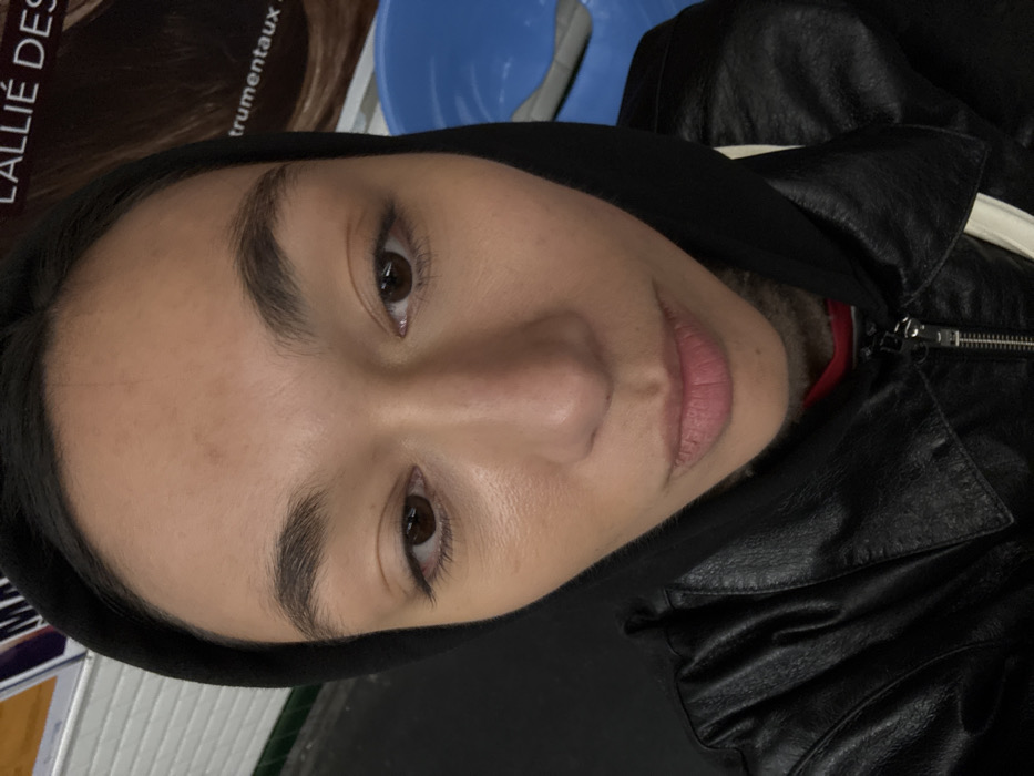 A single individual, appearing to be female, is depicted in a close-up, slightly high-angle portrait, occupying the majority of the frame. She has medium brown skin, dark brown eyes looking slightly downward, dark, full eyebrows, and full lips. Her dark hair is mostly covered by a black hood. She is wearing a black faux leather jacket over a black hooded garment, with a hint of red fabric visible underneath. A white strap, possibly from a bag, is visible over her right shoulder.
The background is blurred, revealing elements consistent with a public transport environment, specifically a station or train interior in Paris, France. To the viewer's left, a dark horizontal surface with a lighter stripe above it is visible, suggestive of a platform edge or interior wall. To the viewer's right, a distinct blue plastic seat or structural element is partially visible, with a light-colored object resting on it. Above the seat, a large advertisement poster displays partial French text, specifically "L'ALLIÉ DES" and "trumentaux", confirming the location as Paris. The poster also shows a section of brown hair, implying an advertisement for hair-related products or services. The scene appears to be dimly lit by ambient sources.