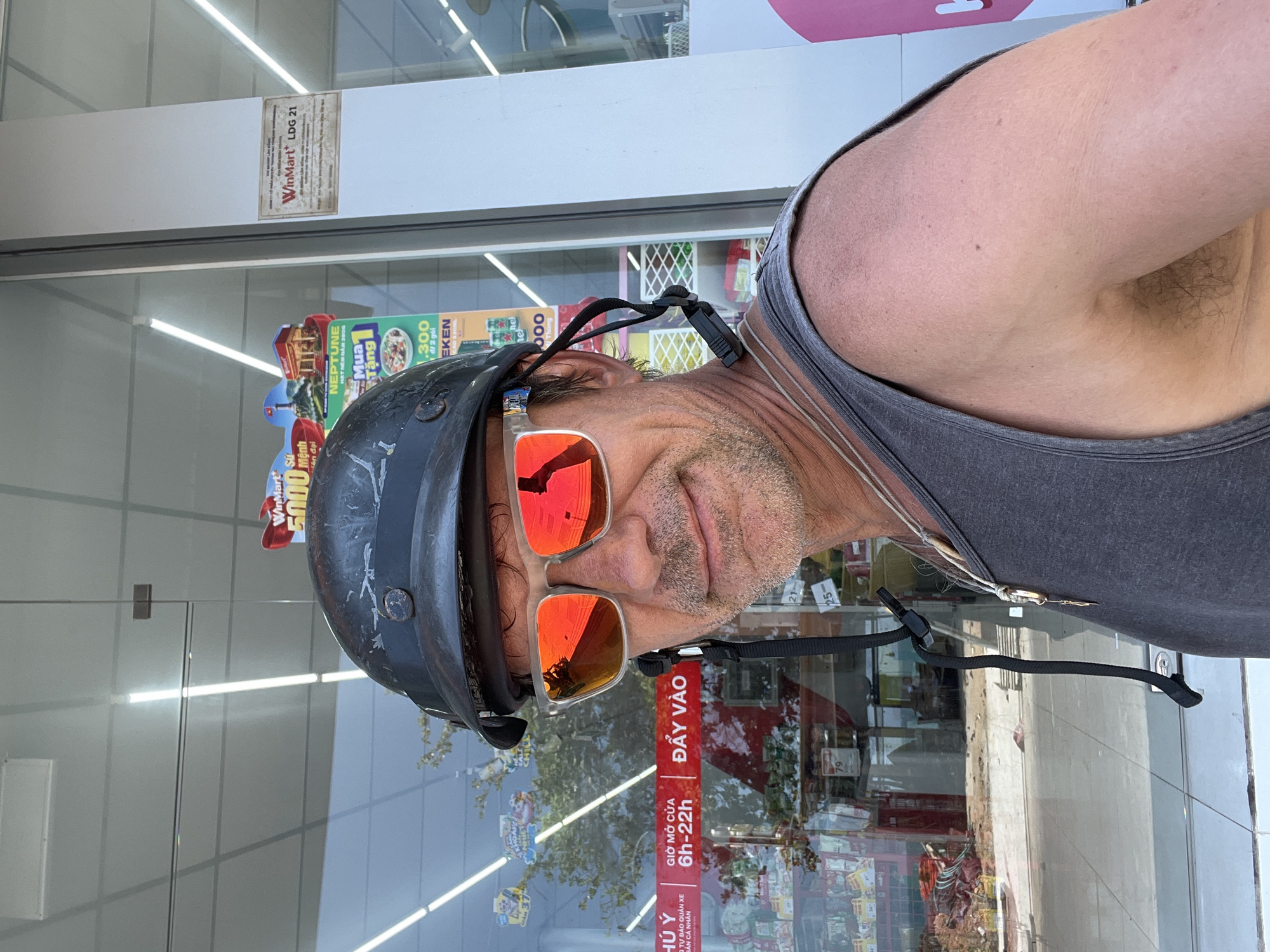 The image shows a man wearing a dark helmet and mirrored sunglasses. He is standing in front of a store with a glass storefront. The store appears to be a convenience store, with shelves of products visible inside. There are signs in Vietnamese on the storefront, indicating the store is likely located in Vietnam. One sign, "WinMart LDG 21," suggests the store is part of the WinMart chain. The man is wearing a gray tank top and has a goatee. The reflection in his sunglasses shows a street scene with buildings and trees. The overall setting suggests an urban or semi-urban environment.