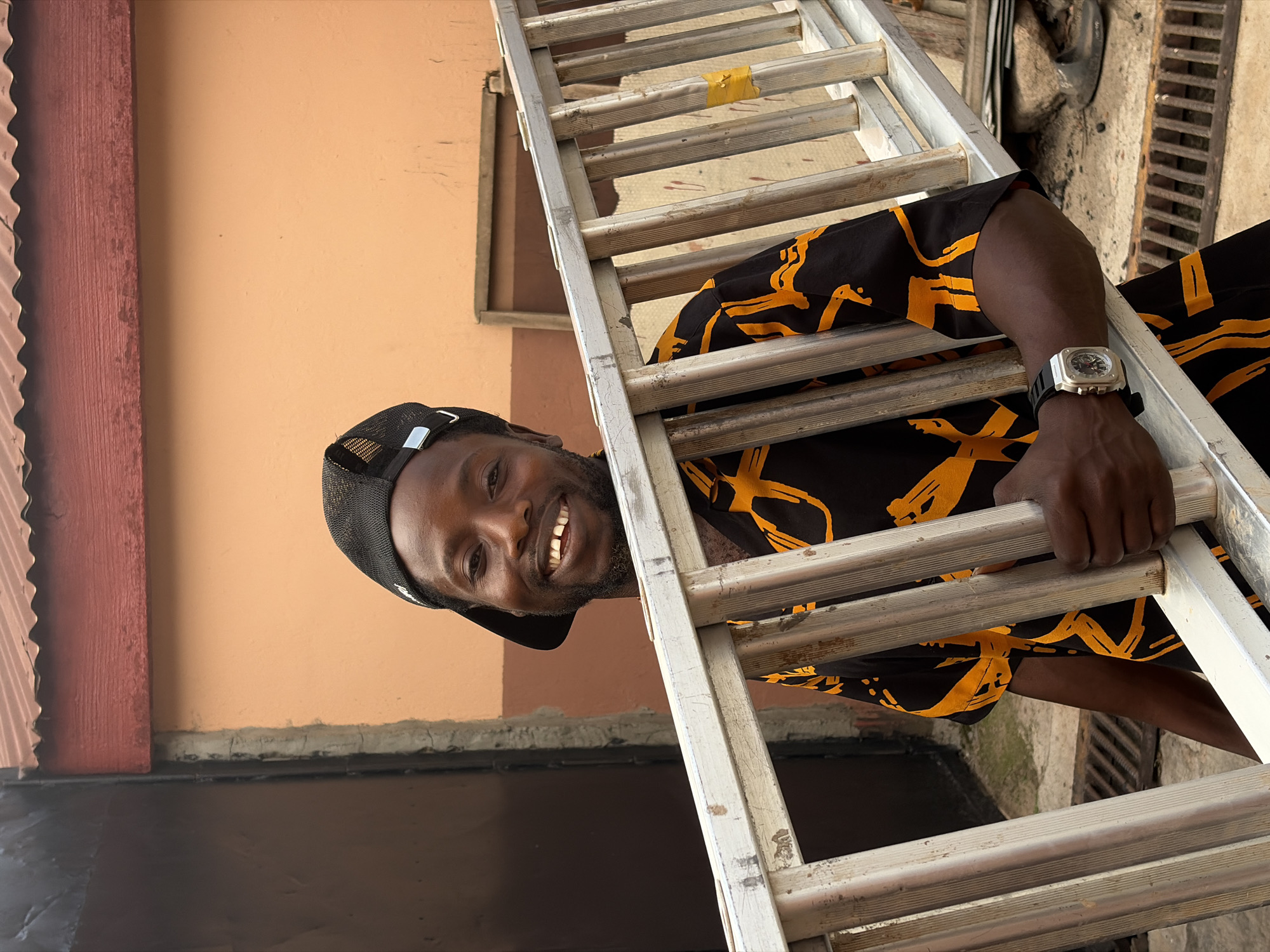A man is seen holding a ladder in what appears to be Jos, Nigeria. The man is wearing a black cap, a black and yellow patterned shirt, and a watch on his left wrist. The ladder is aluminum. The background includes an orange-colored wall and what looks like a drain cover.