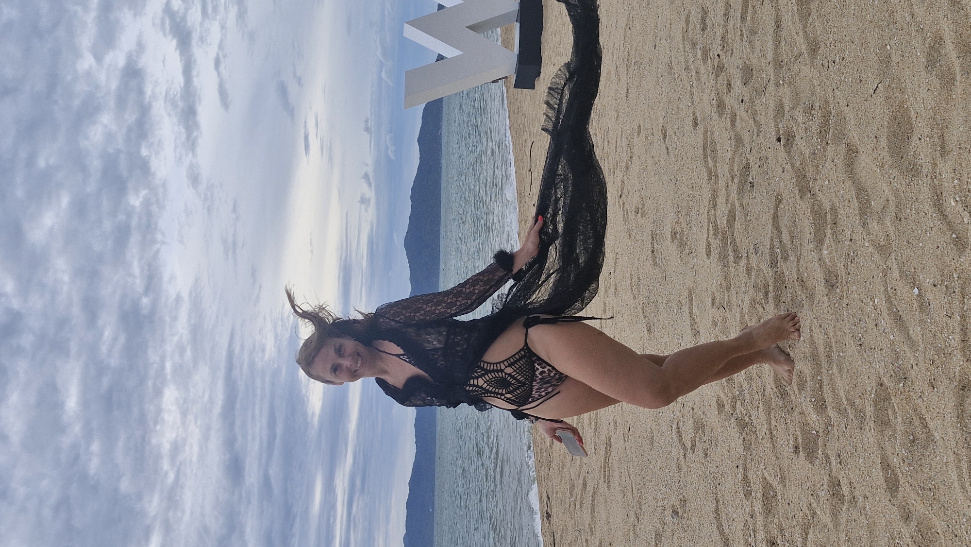 The image shows a woman standing on a sandy beach. She is wearing a black lace cover-up over a leopard print bikini. She is smiling and looking at the camera. Her hair is blowing in the wind. In the background, there is the ocean and a large white letter, possibly part of a sign. The sky is cloudy and overcast. The lighting suggests it is daytime, but the specific time of day is not clear. The overall mood of the image is relaxed and happy.
