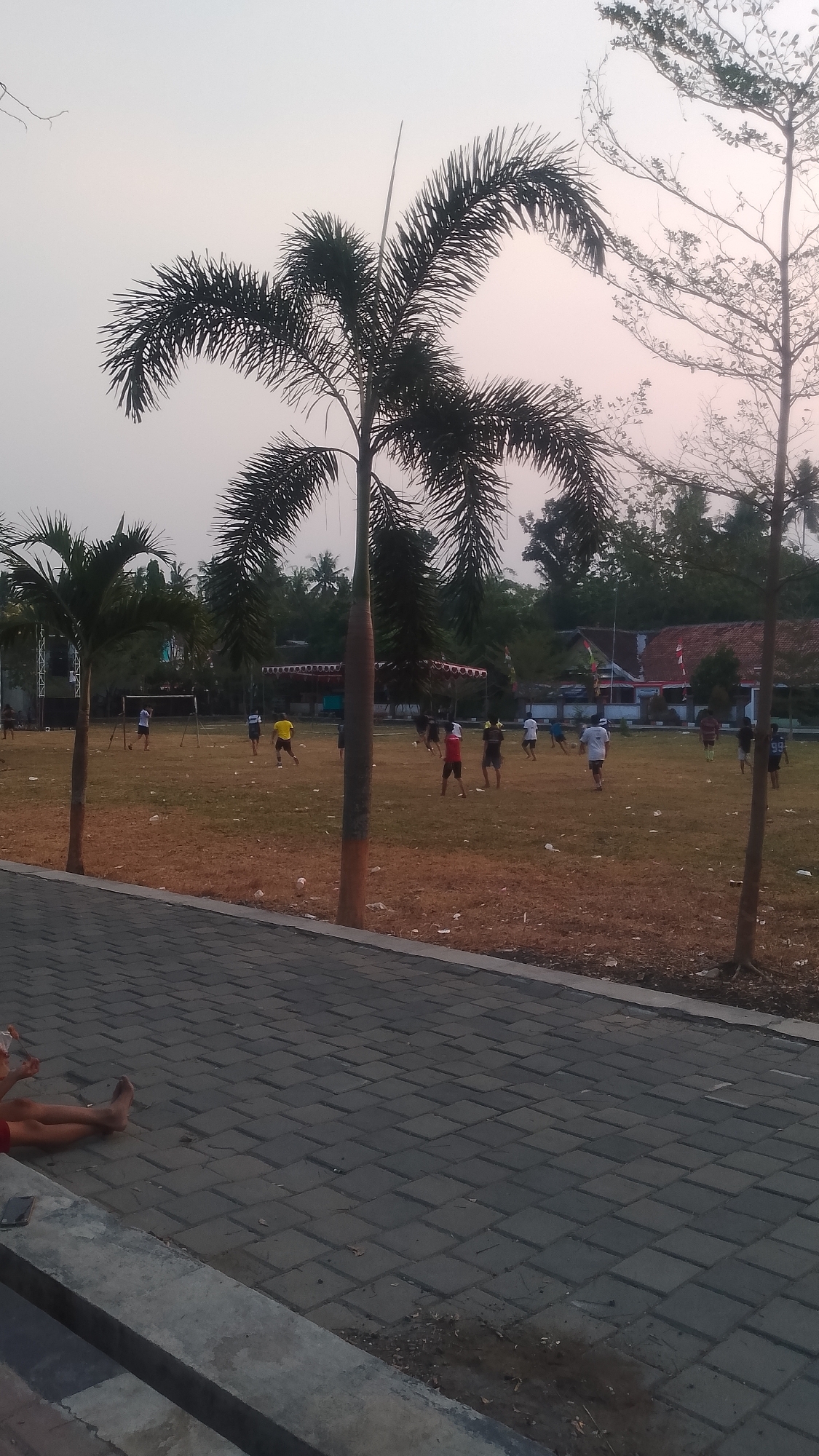 In Karanglangu, Indonesia, a group of individuals are engaged in a soccer game on a dry, grassy field. The game is taking place in the late afternoon or early evening, as indicated by the soft, diffused light. Several palm trees are present in the foreground and midground, with one prominent palm tree in the center of the frame. In the background, there are buildings with red tile roofs and flags, suggesting a village or community area. A paved walkway with a concrete border is in the foreground, where two individuals are seated, one with their legs extended. The sky is overcast with a pale pinkish hue.