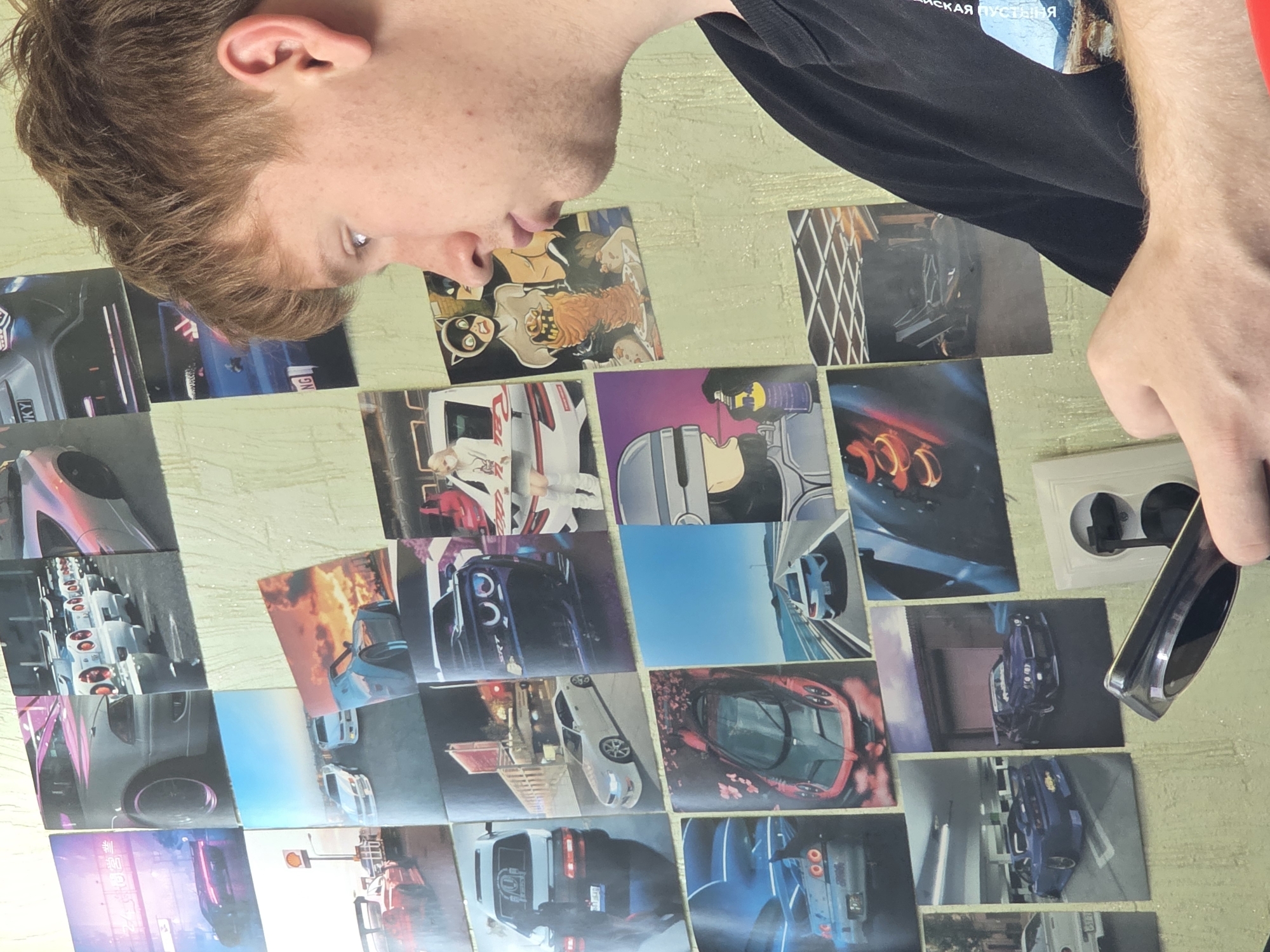The image shows a young man with light brown hair and fair skin, with freckles on his face, in profile, looking to the left. He is wearing a black t-shirt. The background consists of a wall covered with various printed images, primarily featuring cars. The images include a variety of car models, some in action shots, others posed. Some of the images are anime-style illustrations or posters. A white electrical outlet and a silver object, possibly a phone, are visible in the foreground, obscuring part of the lower section of the wall. The overall impression is a person in a room decorated with car-themed posters. No specific location in the city of Chita, Russia, can be identified from the provided image.