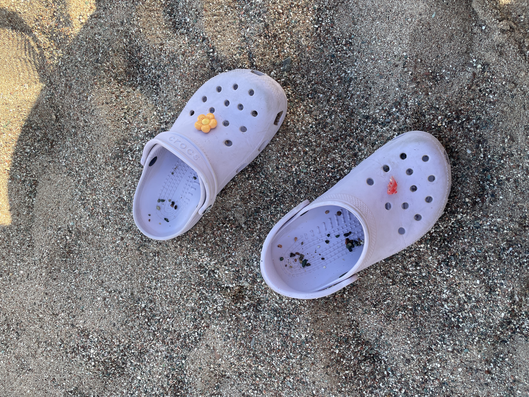 Two light purple Crocs are visible on a beach of small pebbles and sand. One Croc, positioned in the upper right of the frame, has a small orange flower charm attached. The other Croc, in the lower left, has a small red object embedded in it. The pebbles on the beach are varied in color, including shades of grey, white, and brown. A shadow falls across the upper right portion of the sand, suggesting a sunny day. The setting appears to be the beach in Rafailovići, Montenegro.