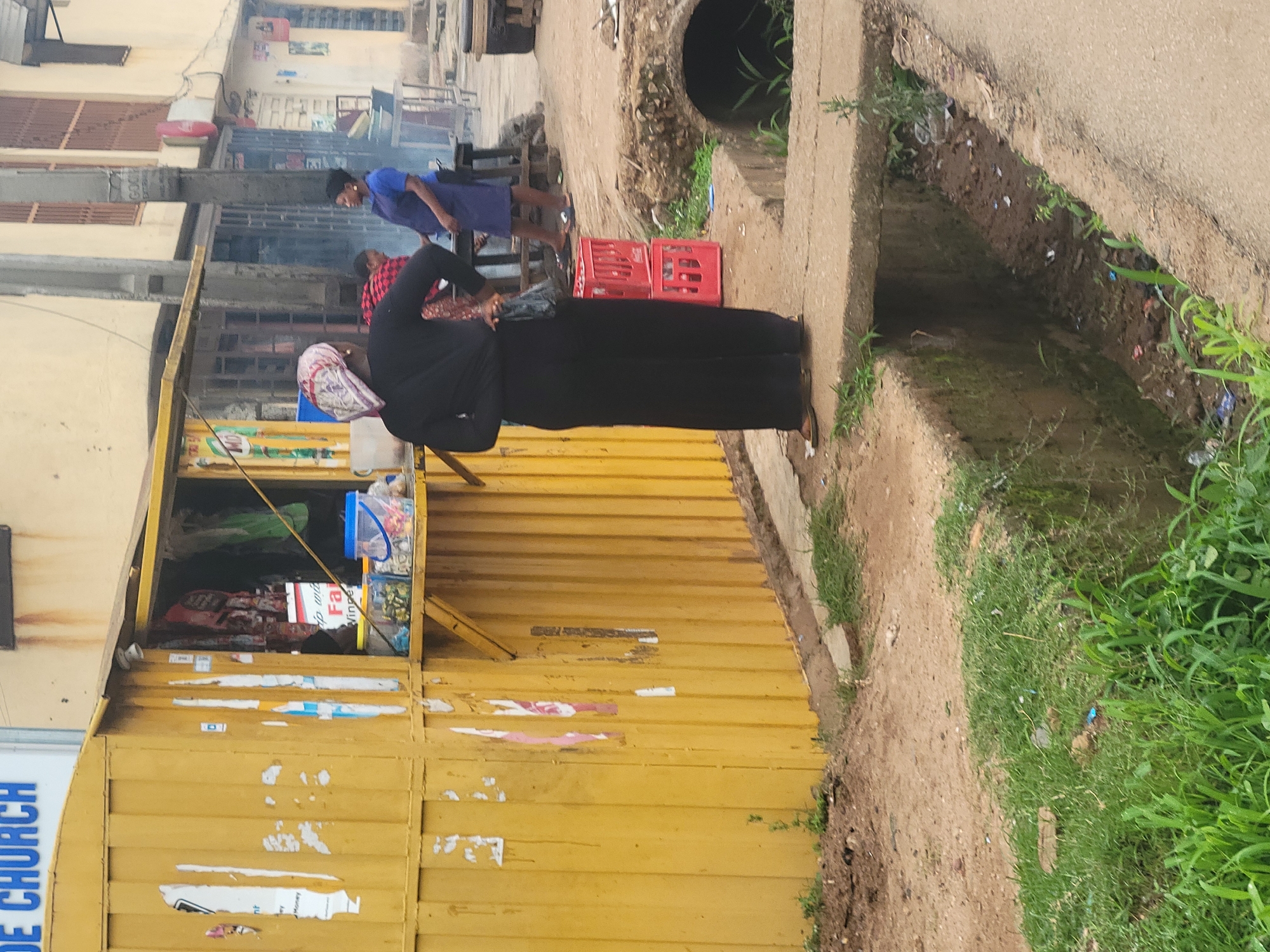 In the city of Unknown, Unknown, a scene unfolds at what appears to be a street-side vendor. A woman, dressed in a black long-sleeved top and wide-legged black pants, stands facing away from the camera, her left hand on her hip. She wears a patterned headscarf and flip-flops. To her right, another woman in a blue dress and black head covering is partially visible, appearing to interact with something at a stall. Behind the first woman, red plastic crates are stacked.
The vendor stall itself is a bright yellow corrugated metal structure, open at the front. Shelves within display various items, including what appear to be packaged goods and colorful buckets. The stall is adorned with peeling posters and signs. In the foreground, a concrete path runs alongside a drainage ditch filled with murky water and debris. Green grass lines the edge of the path and the ditch. The background reveals more of the urban environment with buildings and other structures, suggesting a busy, possibly informal, commercial area. The overall atmosphere is one of everyday street life in an outdoor market setting.