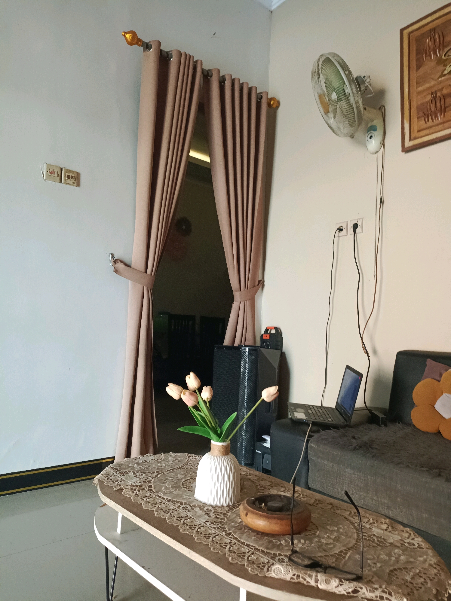 The image depicts an interior living room scene, likely in Rancang Kencono, Indonesia. The room features a light-colored wall with electrical outlets and light switches. A brown curtain hangs from a gold-capped rod, partially obscuring a doorway. A wall-mounted fan is visible on the right side of the wall, with its cord plugged into an outlet. A framed piece of artwork hangs to the right of the fan. In the foreground, a coffee table is set with a lace doily, a white vase containing several light pink tulips, a wooden ashtray with a burnt cigarette butt, and a pair of eyeglasses. Behind the coffee table, a dark-colored sofa is partially visible, with a patterned cushion. A laptop computer rests on a dark surface to the left of the sofa, and a black speaker system is positioned behind the coffee table. The lighting suggests it is daytime.