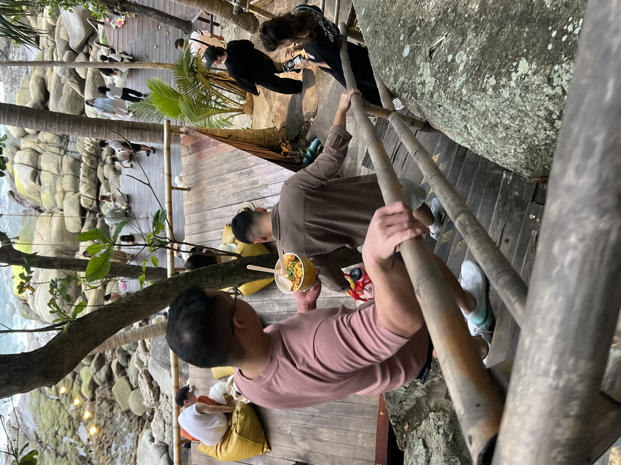 The image depicts an outdoor scene in Karon, Thailand, featuring a wooden multi-level deck overlooking a rocky shoreline with the ocean in the background. In the foreground, a person in a pink t-shirt is descending wooden stairs, holding a bowl of food. To their right and slightly behind, another person in a brown shirt is also on the stairs, facing away from the camera. Further up the stairs, a person in a black outfit is looking at their phone. Several other individuals are visible on the upper deck, some sitting on yellow beanbags, while others are standing or walking. The environment includes tropical trees and plants, large rocks, and decorative string lights. The overall atmosphere appears to be a casual gathering or dining area in a scenic, natural setting.