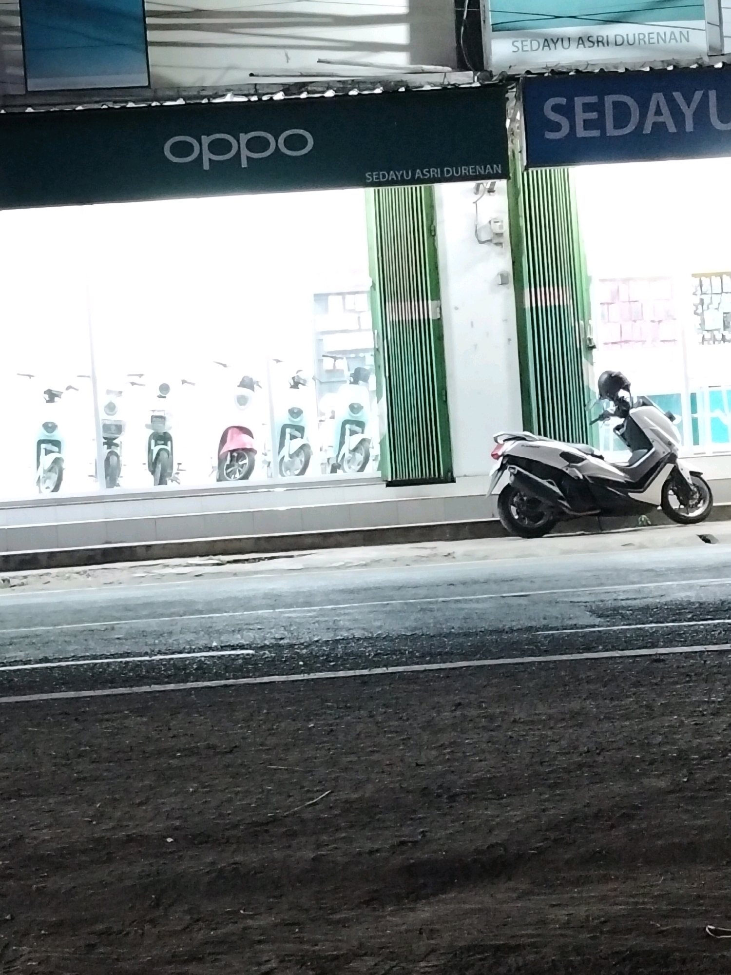 The image depicts a street view in Durenan, Indonesia, likely in front of a commercial establishment. On the left, a sign displays the logo "oppo" in white letters against a dark green background. To the right of the "oppo" sign, a blue sign reads "SEDAYU ASRI DURENAN" in white letters. Behind these signs, a store window displays several white scooters. In the foreground on the right, a white and black scooter with a black helmet on its handlebars is parked on the side of the road. The road itself is visible in the lower portion of the image, appearing wet and dark. The overall scene suggests a business district at dusk or night, with artificial lighting illuminating the storefront and vehicles.