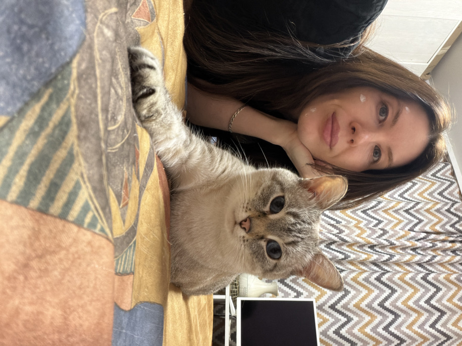 A woman with long brown hair rests her head on her hand, posing next to a light brown tabby cat. The cat lies with its paw extended, looking directly at the viewer. The backdrop consists of a patterned bedsheet in hues of blue, yellow, and brown, and a zigzag patterned curtain. A black laptop screen is partially visible in the bottom right corner.