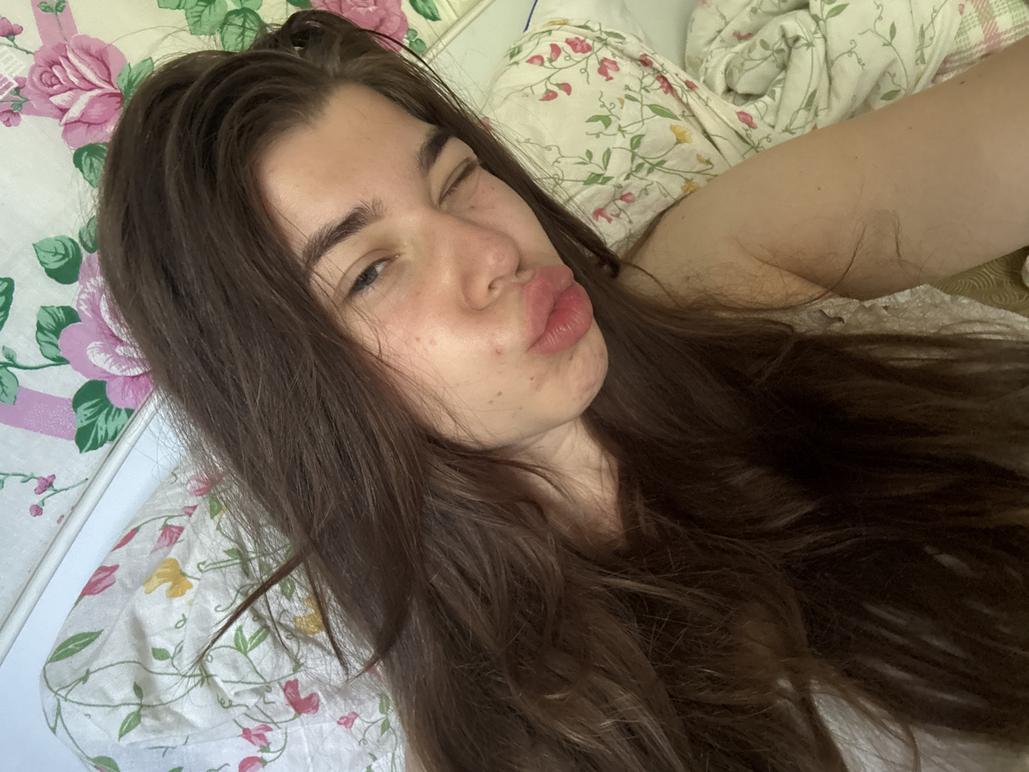The image shows a young woman with long, dark brown hair lying in bed. She is looking at the camera with one eye closed in a playful wink and her lips puckered. Her skin appears slightly flushed, and she has a few blemishes on her face. The background is a white bed with floral-patterned bedding in shades of pink, green, and yellow. The overall impression is of a casual, at-home setting. There is no indication of the specific location within Мещёры, Russia.