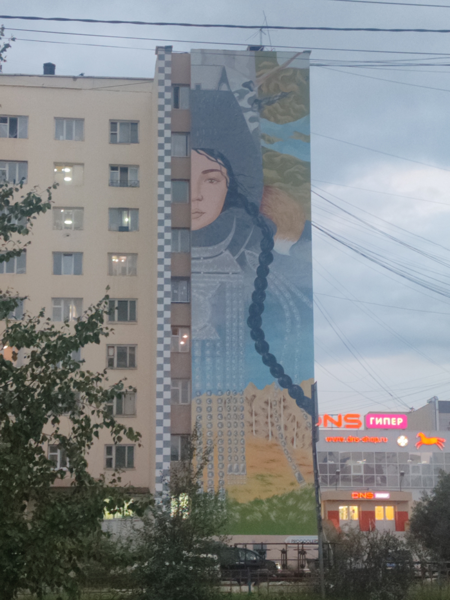 The image shows a mural on the side of a tall apartment building in Yakutsk, Russia. The mural depicts a woman's face with a long braid, and is surrounded by abstract designs and landscapes. The building is light yellow with multiple floors and windows. To the right of the apartment building, there is a commercial building with a neon sign that reads "DNS" and "ГИПЕР". The sky is overcast. The image appears to have been taken from street level, with trees and foliage in the foreground.