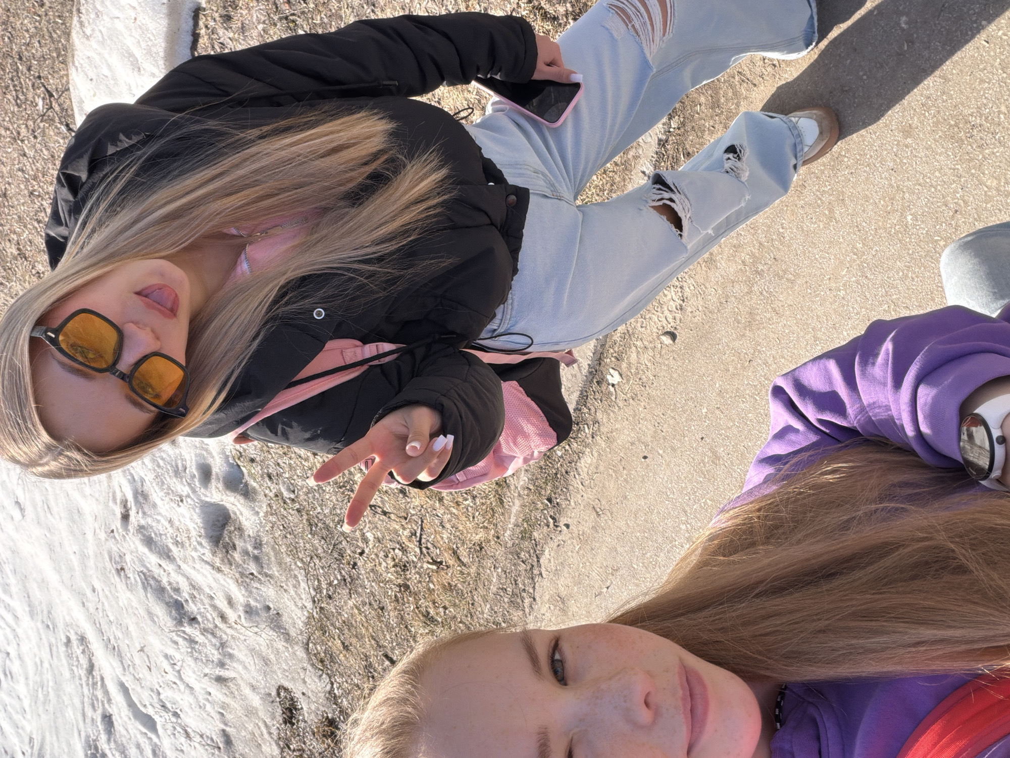 Two young women are visible in a selfie. One woman wears a black puffer jacket with pink lining, light blue ripped jeans, and brown shoes. She wears orange-tinted sunglasses and displays a peace sign. The second woman wears a purple hoodie. The location appears to be an outdoor area possibly in Berezniki, Russia, given the presence of snow on the ground. The background consists of a mix of snow and concrete.